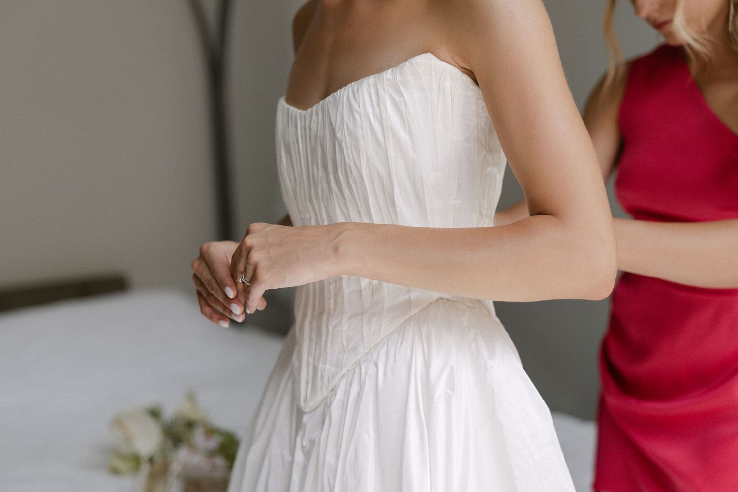 Bridesmaid in red dress helping bride fasten her white strapless wedding gown before the ceremony.