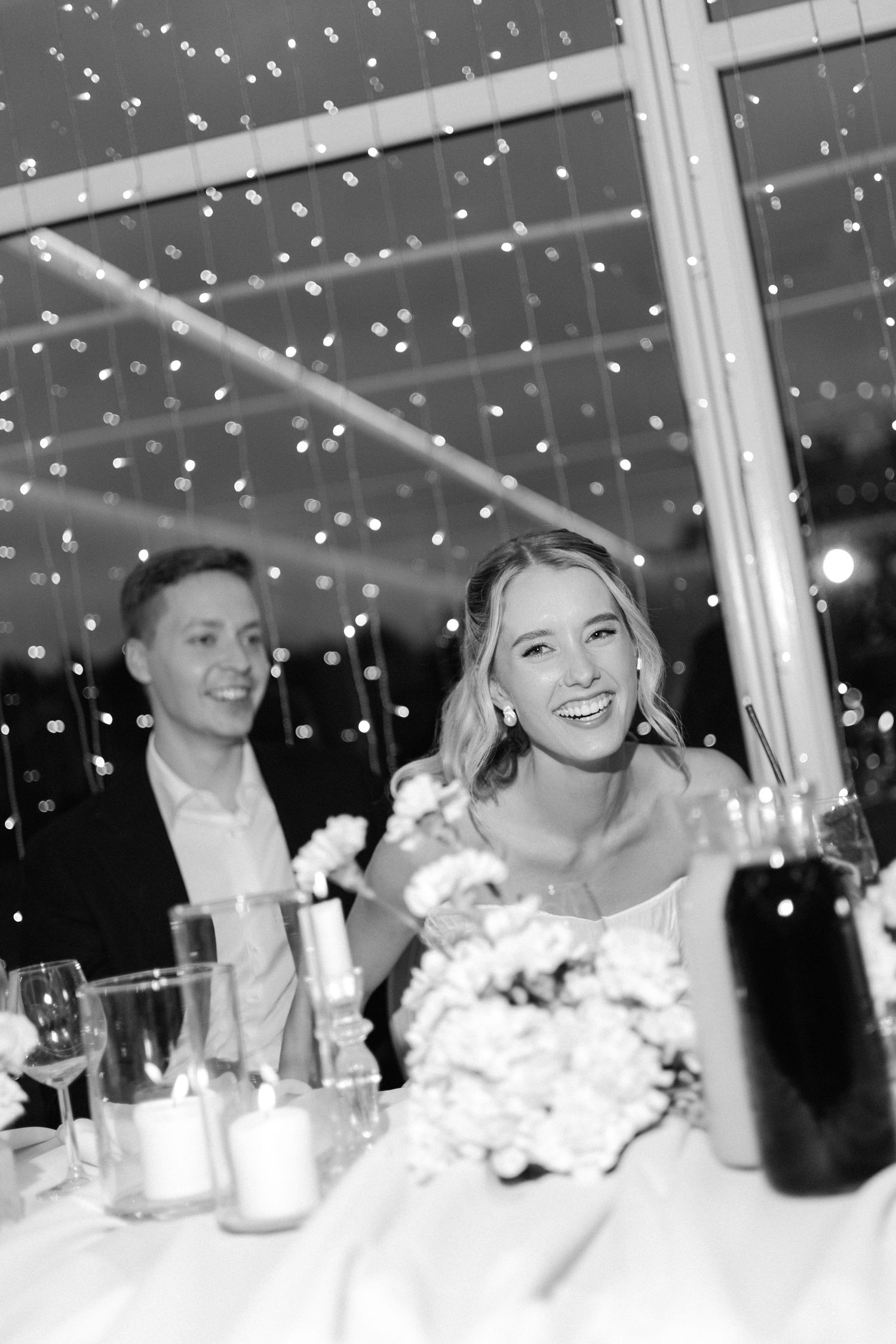 Black and white photo of laughing bride and groom at reception table under twinkling fairy lights.