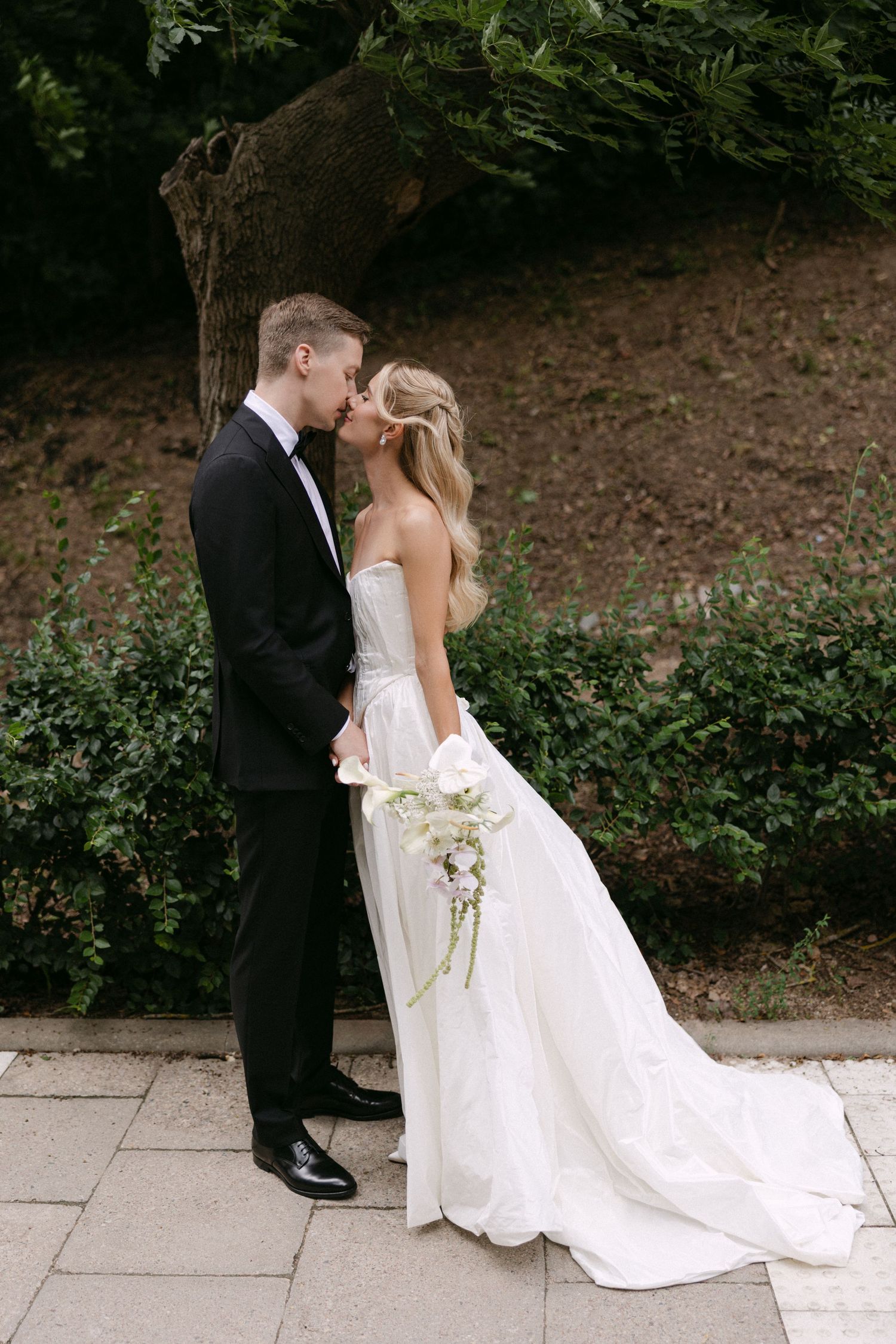 Bride and groom share a romantic kiss outdoors, surrounded by lush green foliage on their wedding day.