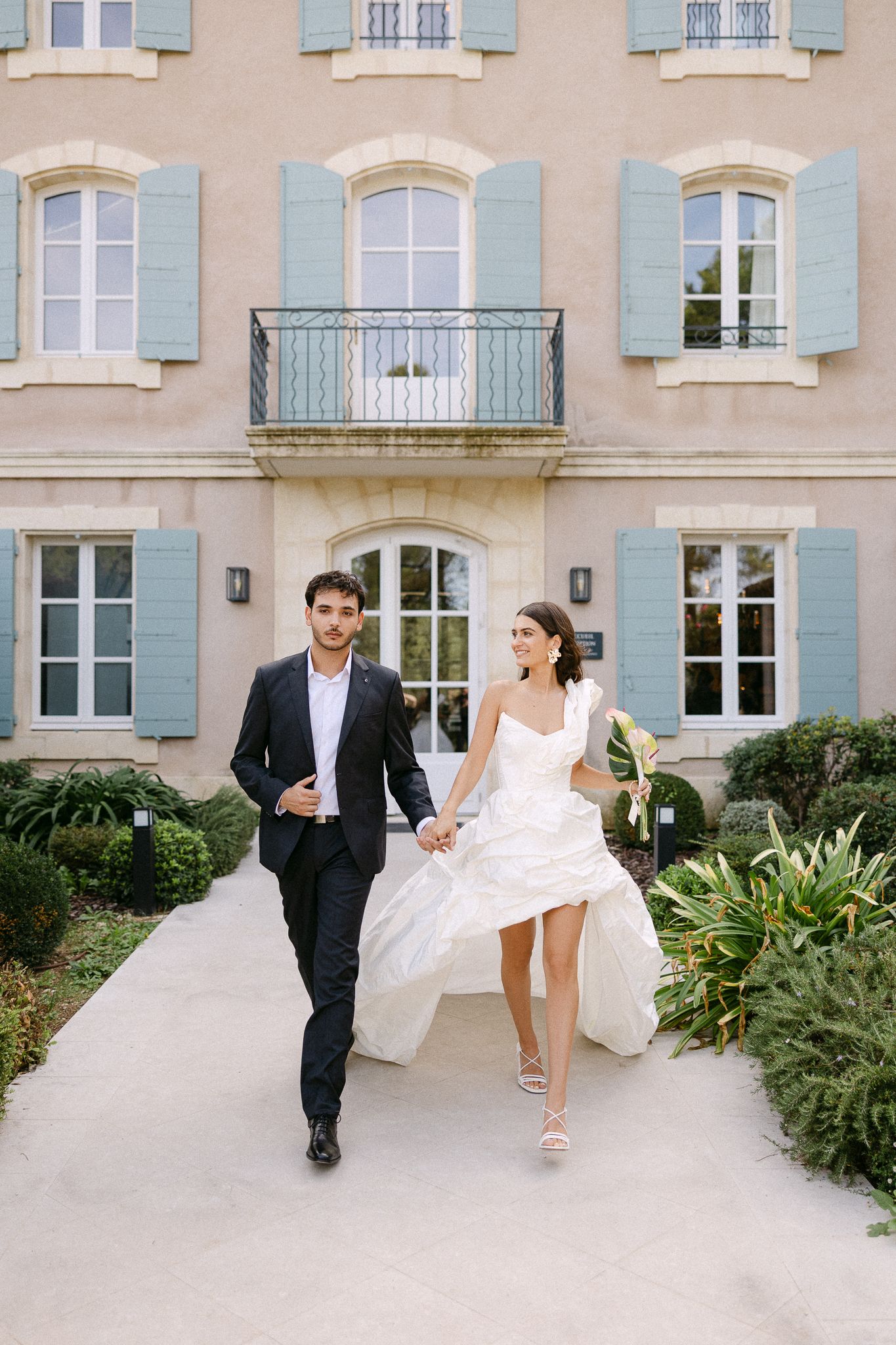 Bride and groom walk hand-in-hand toward camera outside elegant French villa with blue shutters.