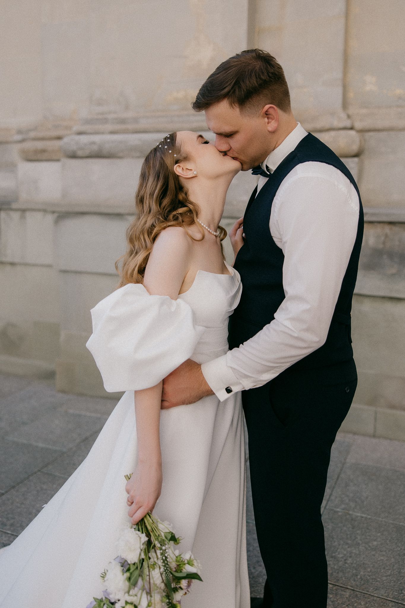 Romantic wedding couple sharing a kiss against a stone building backdrop.