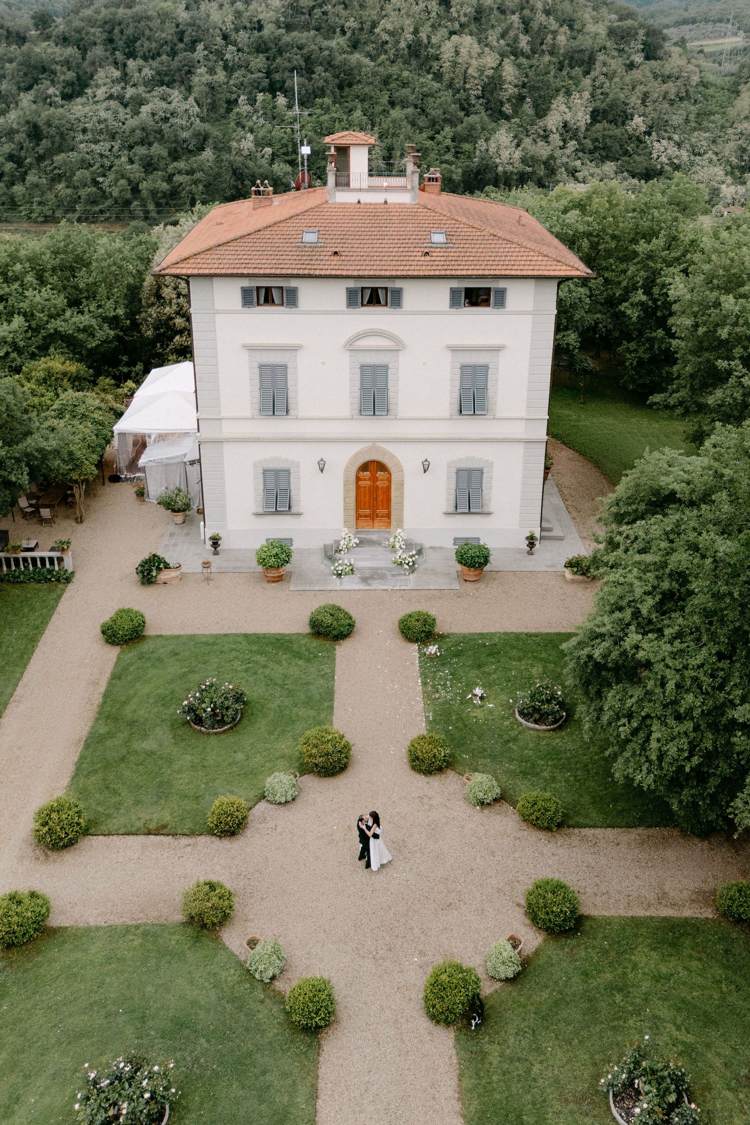 Aerial drone view of newlyweds embracing in geometric garden of white Italian villa surrounded by hills.