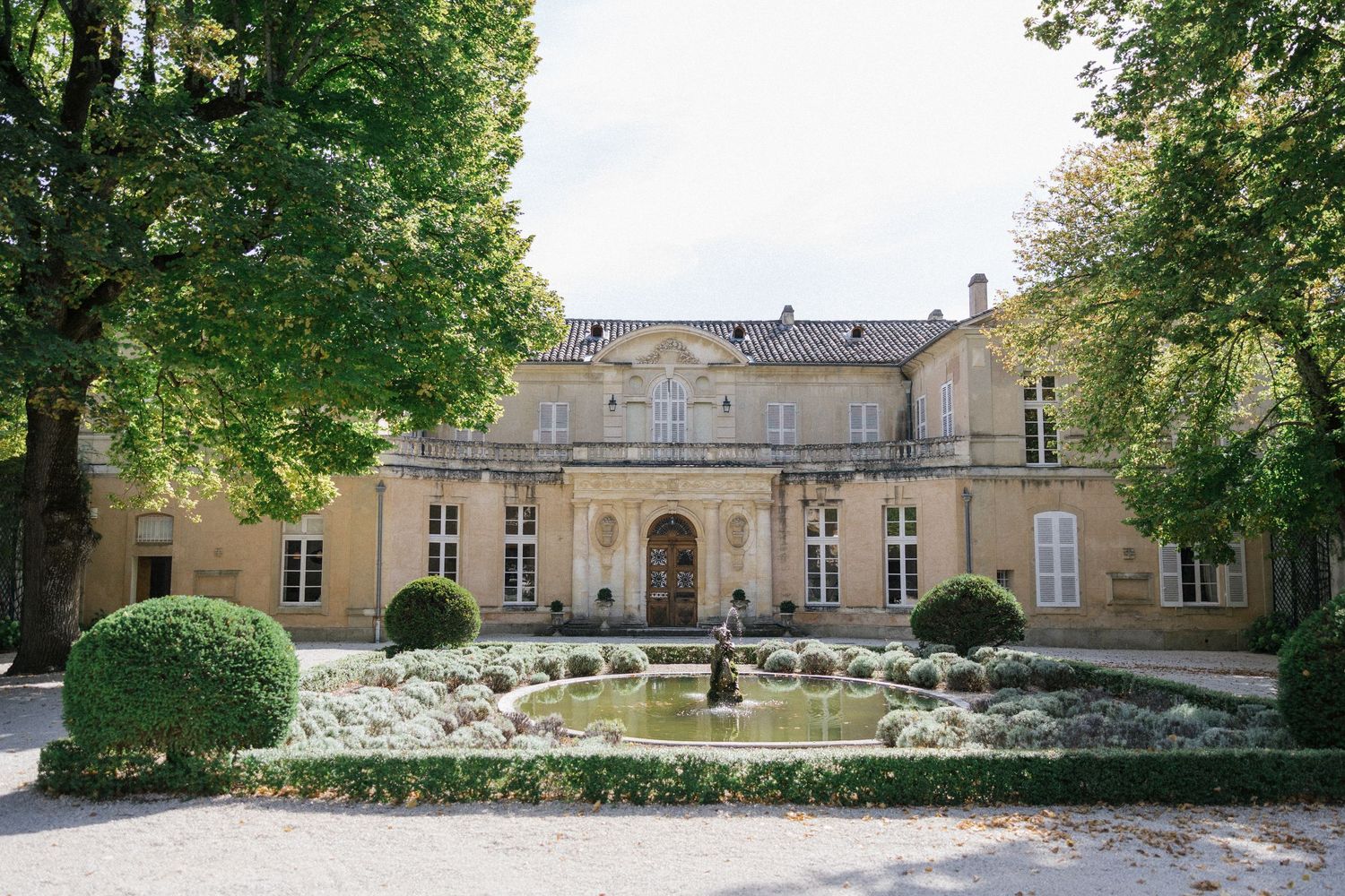 Elegant French chateau with fountain, manicured hedges, and tree-lined courtyard in golden stone architecture.