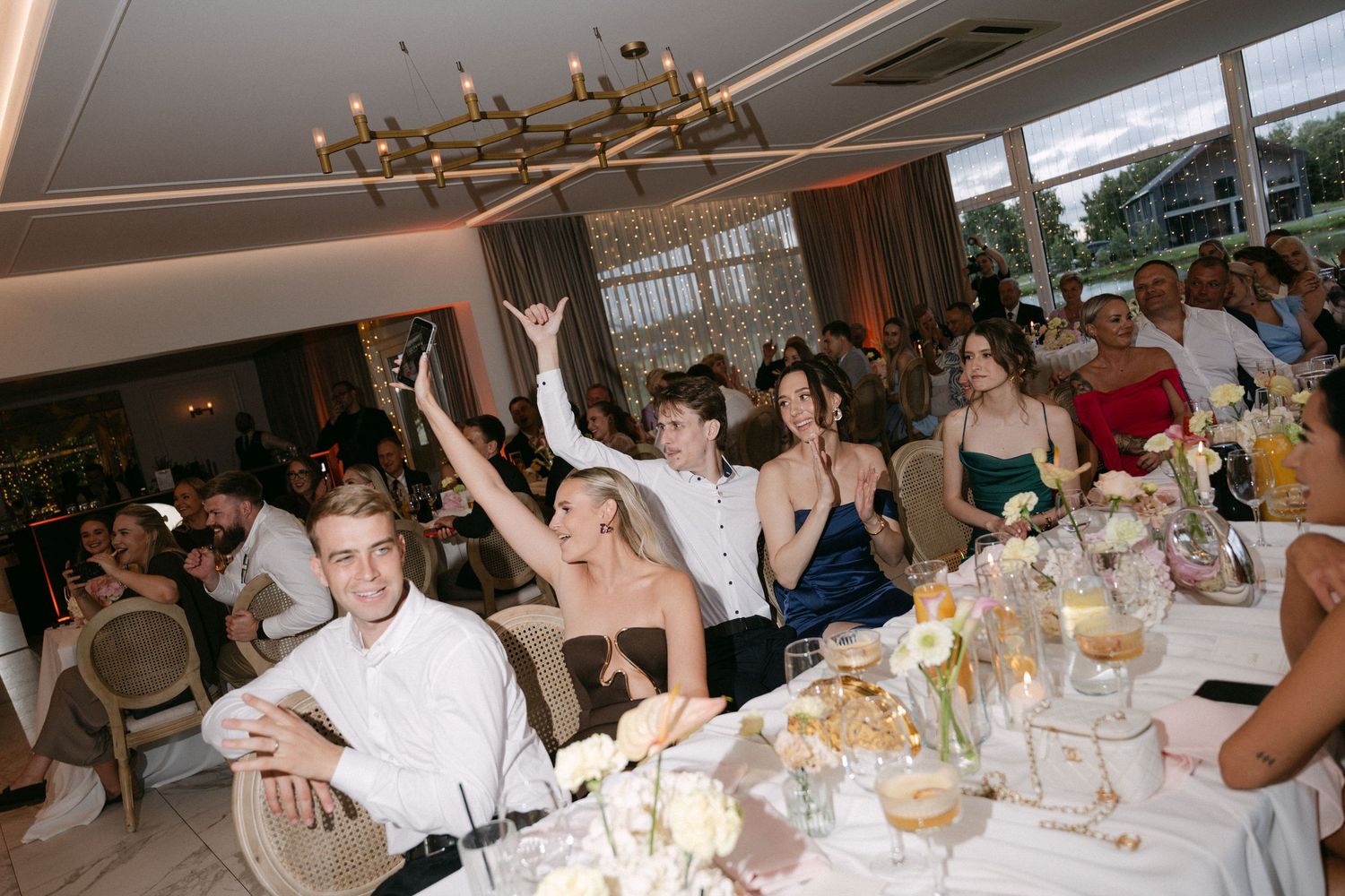 Lively wedding reception with guests celebrating at a decorated table under a gold chandelier.