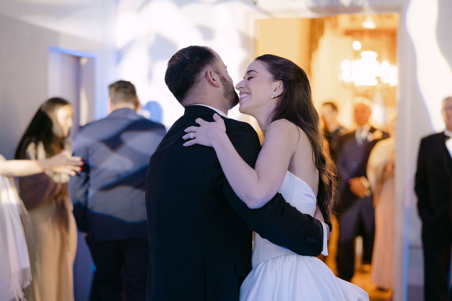 Bride and groom share joyful first dance embrace surrounded by guests at elegant wedding reception.