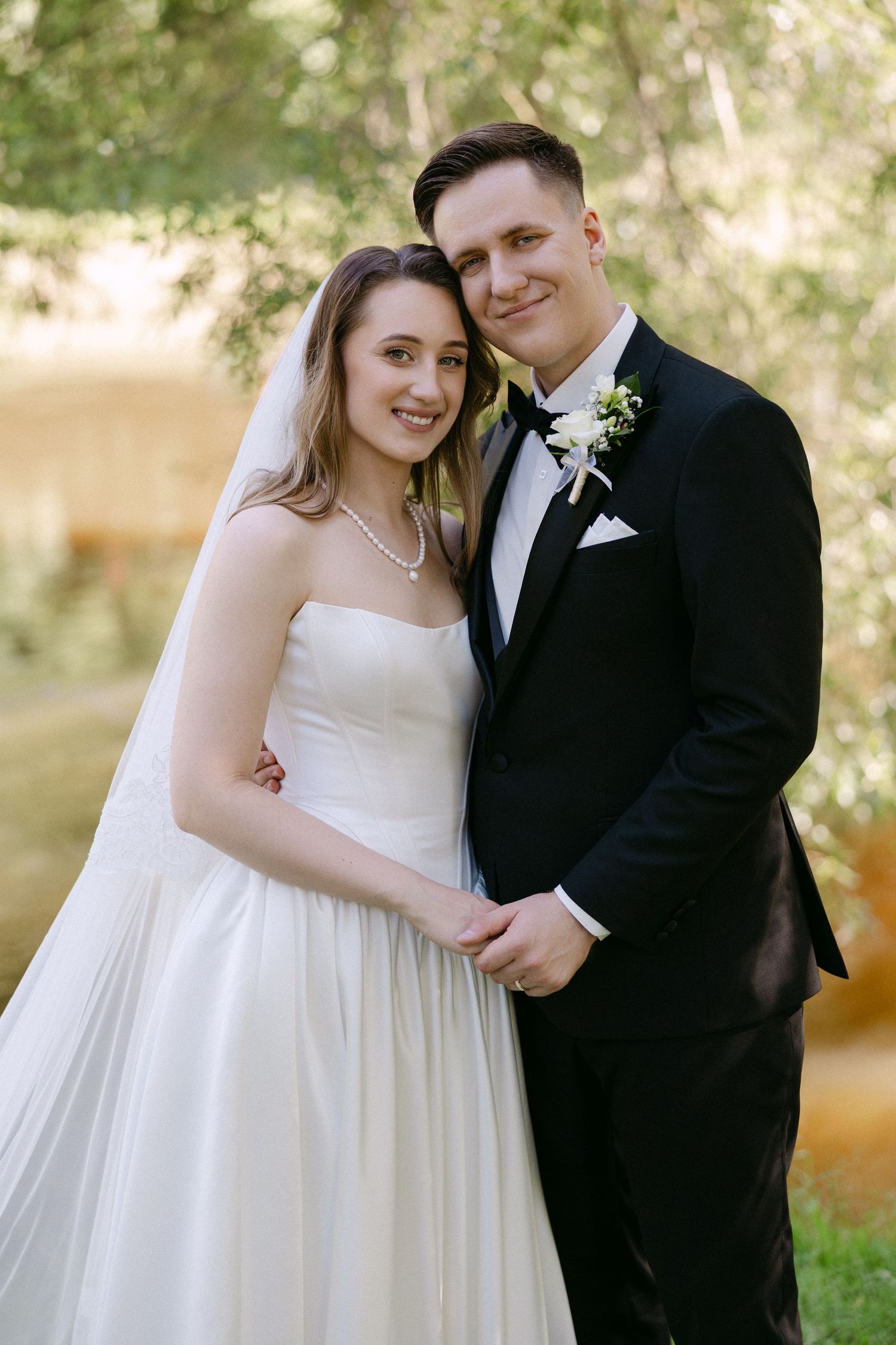 Bride in strapless white gown and groom in black tuxedo pose together outdoors on their wedding day.