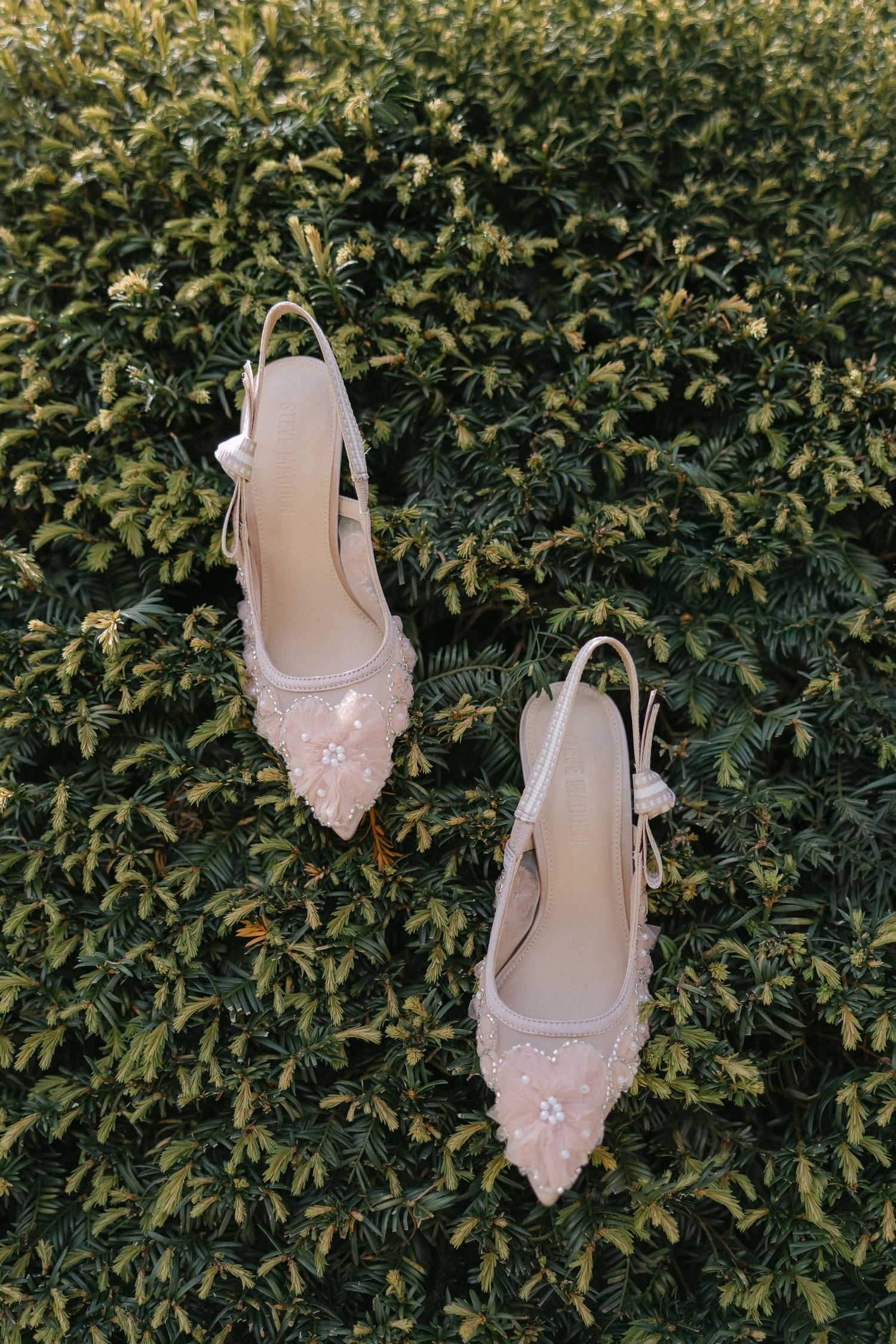 Elegant blush slingback heels with floral embellishments resting on a lush green hedge for a wedding.