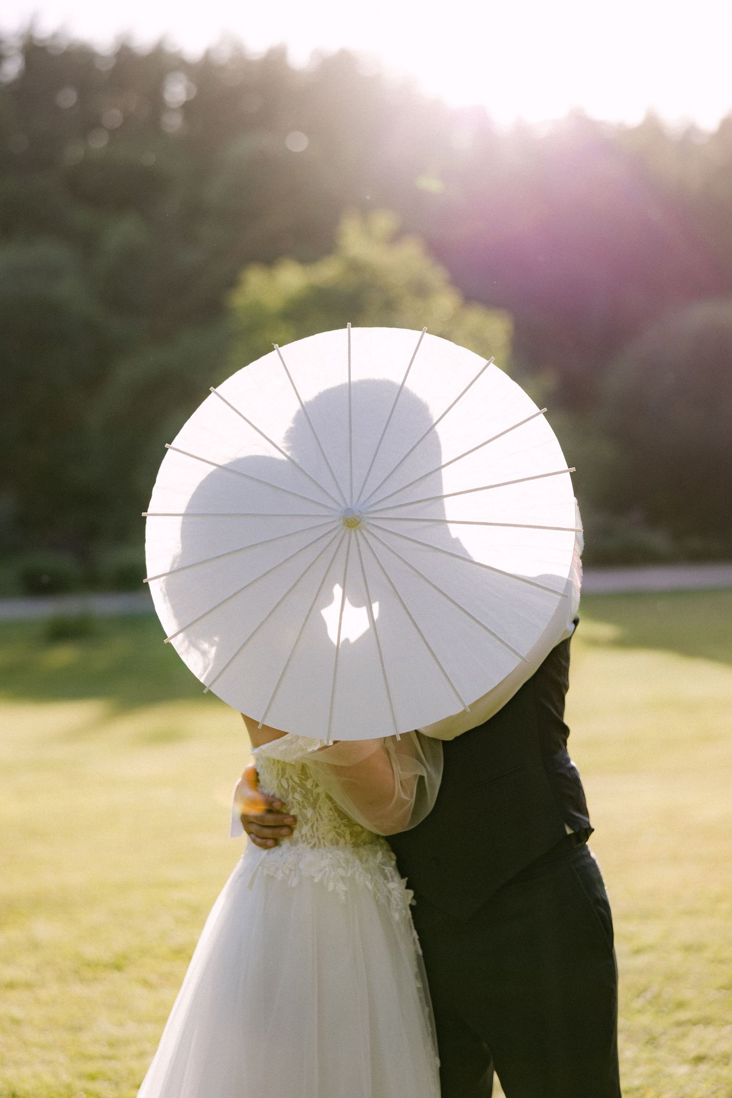 Bride and groom kissing behind a white parasol umbrella in a sunlit outdoor wedding setting.