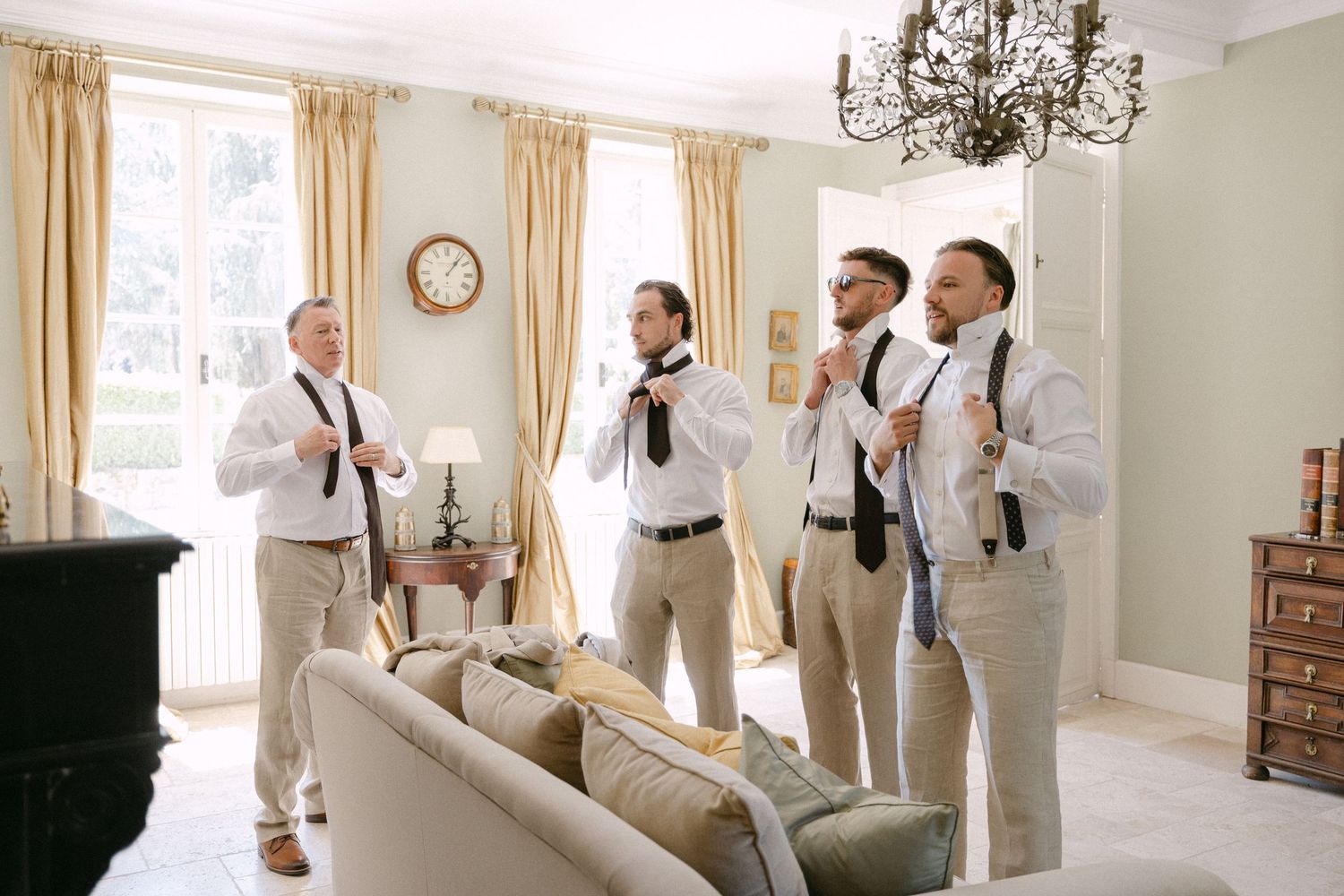 Four groomsmen in white shirts and khaki pants adjusting their ties in an elegant room before a wedding ceremony.