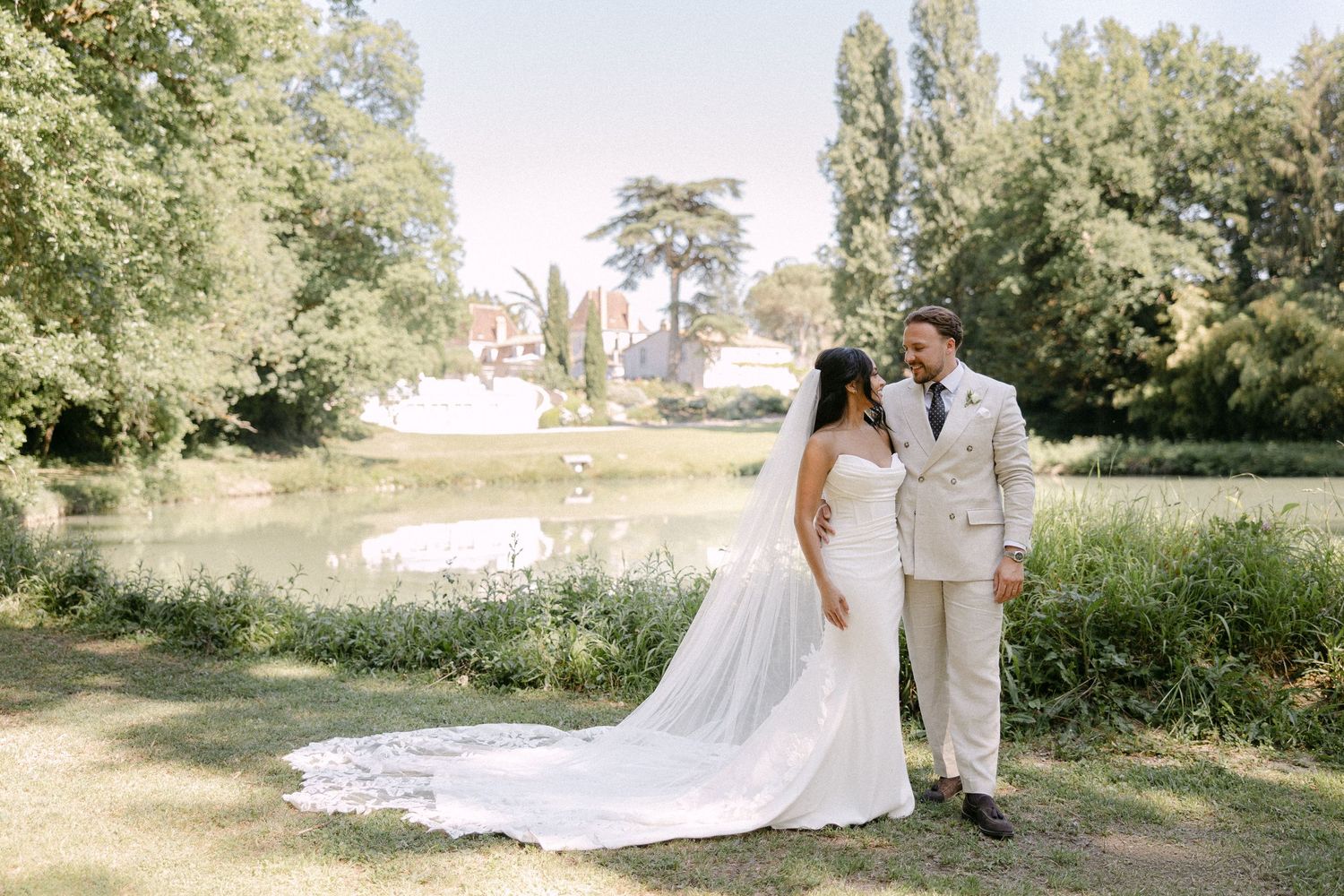 Bride and groom holding hands by a serene lake at a lush French chateau wedding.