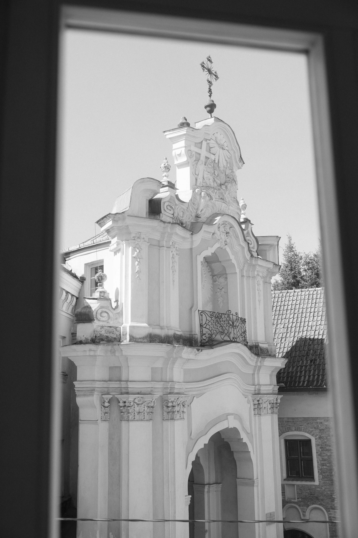 Black and white photo of ornate Baroque church facade with cross, viewed through a window frame.