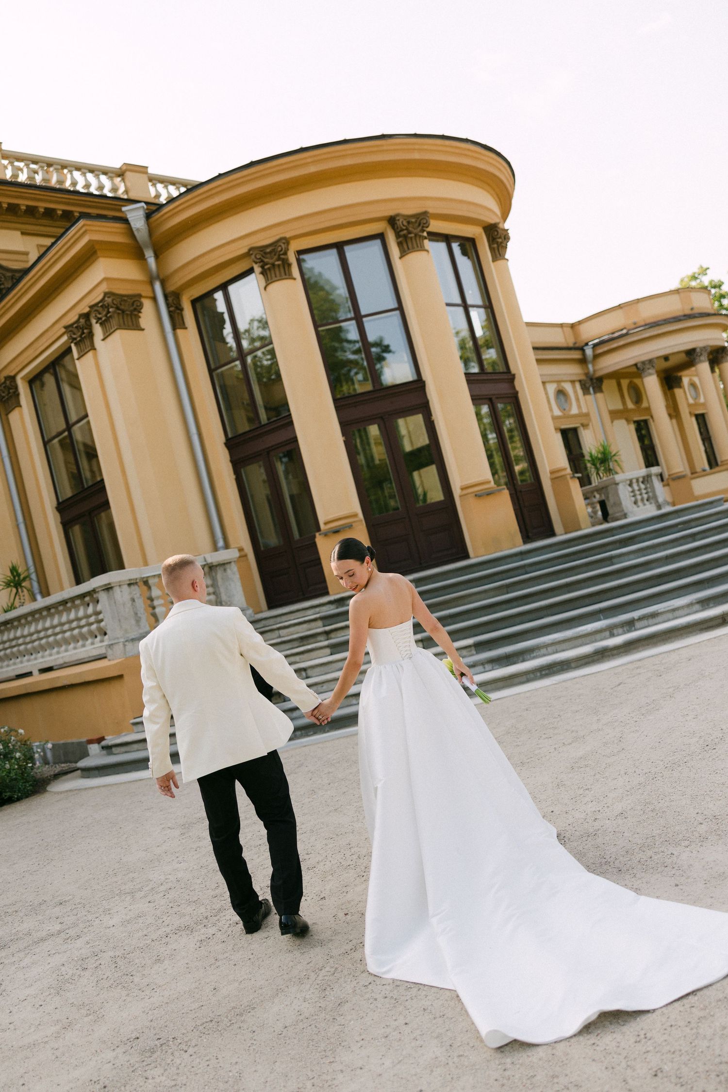 Bride and groom holding hands walking toward a grand neoclassical building on their wedding day.