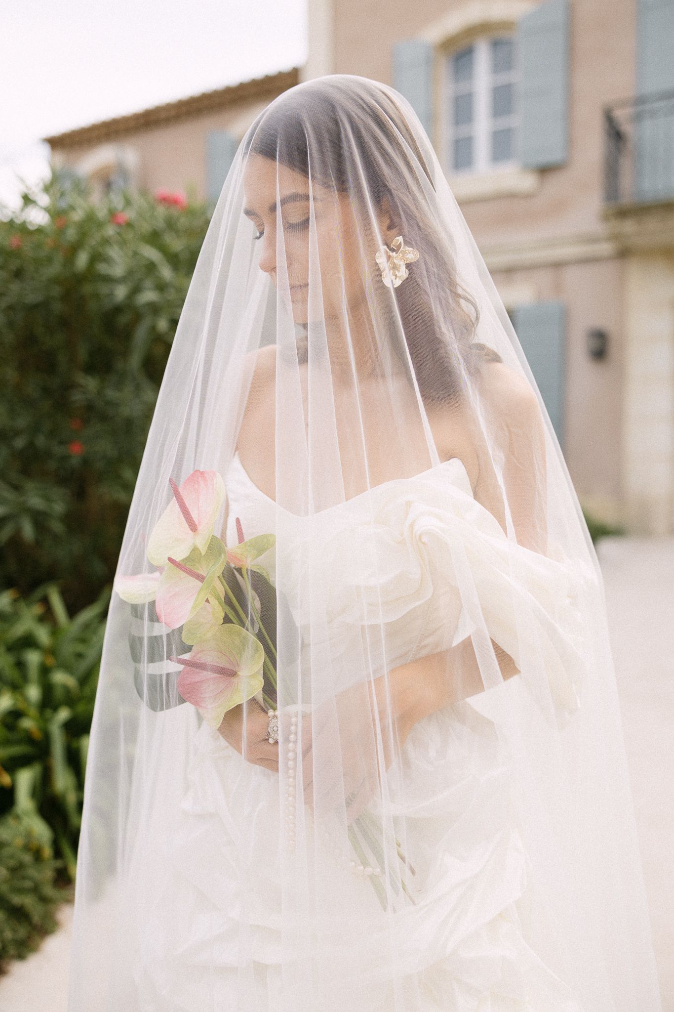 Bride draped in flowing veil holding bouquet of white and pink flowers outdoors.