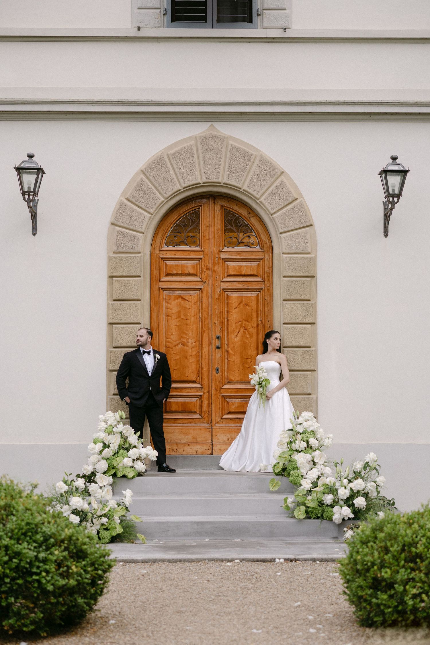 Bride and groom posing on steps of elegant Italian villa with white floral arrangements flanking wooden door.