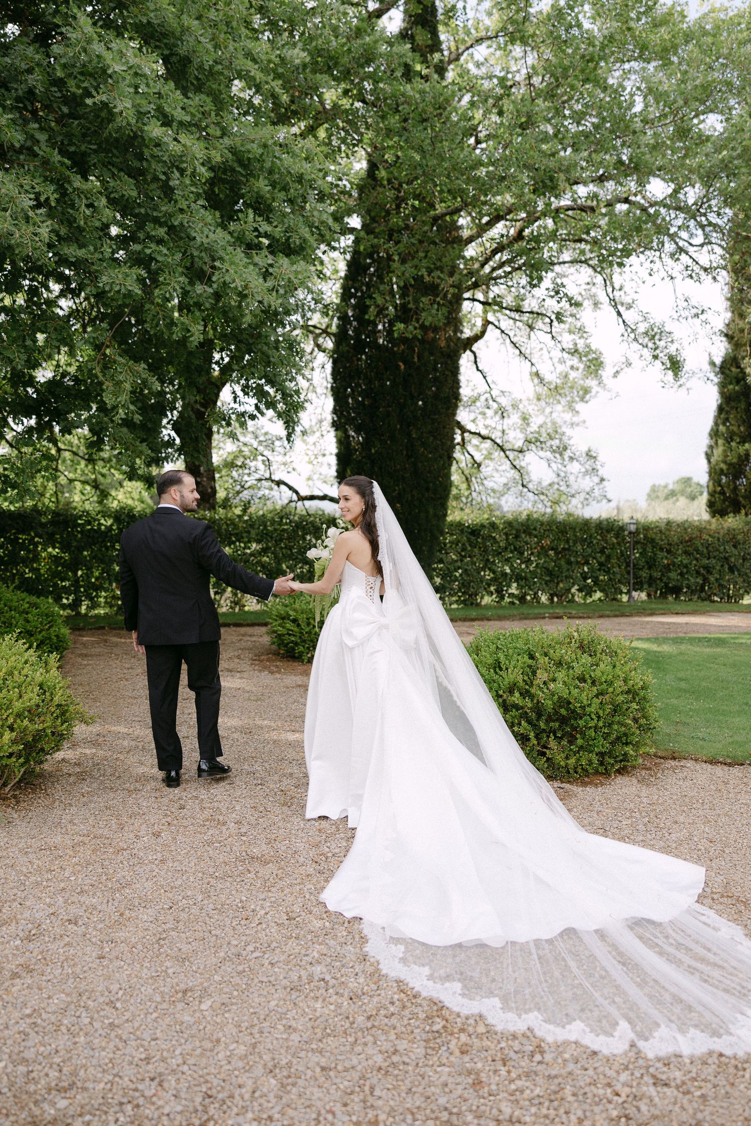 Bride and groom holding hands in lush garden, bride wearing long lace cathedral veil trailing on gravel path.