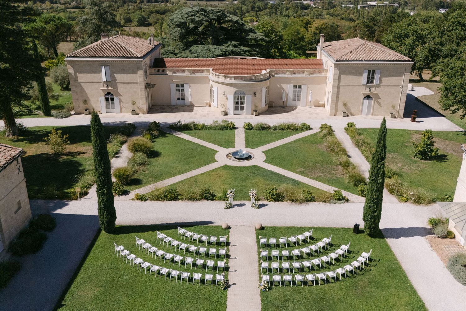 Aerial view of elegant French château with formal garden, fountain, and white chairs arranged for outdoor wedding ceremony.