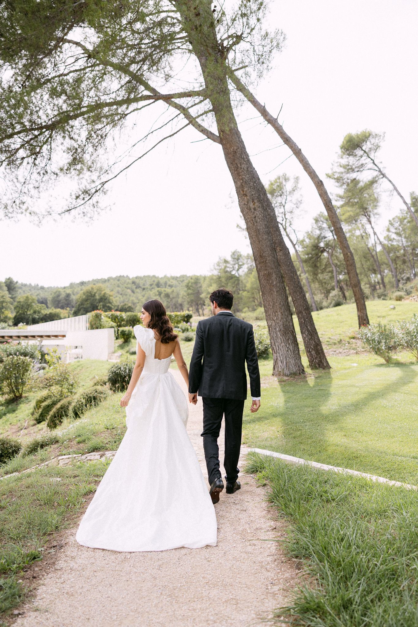 Bride and groom walking hand in hand along garden path lined with tall pine trees.