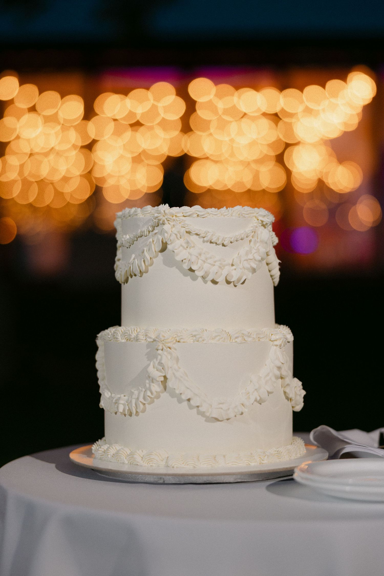 Elegant two-tier white wedding cake with piped swag frosting details on a draped table with bokeh lights in background.