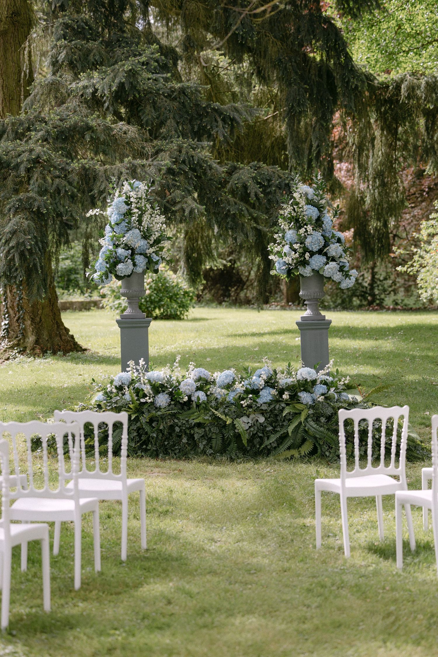 Elegant outdoor wedding ceremony setup with white chairs and blue floral arrangements on lush green lawn.