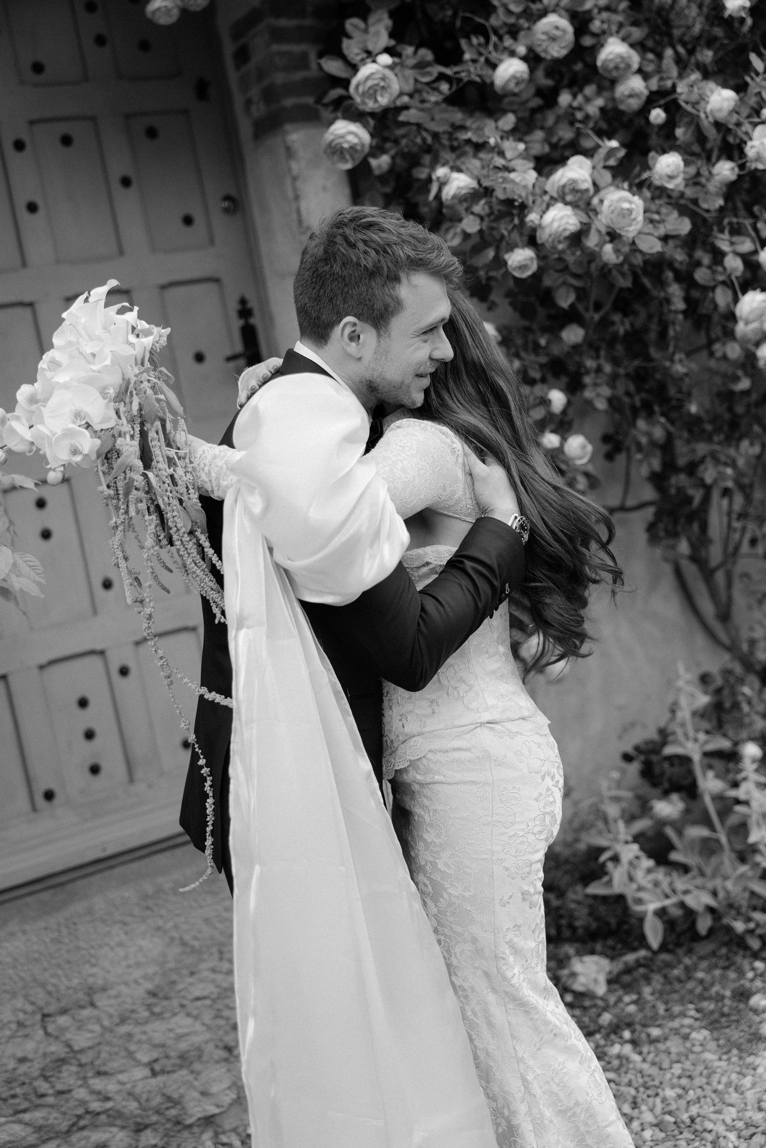Black and white photo of bride and groom embracing near floral-covered door after wedding ceremony.