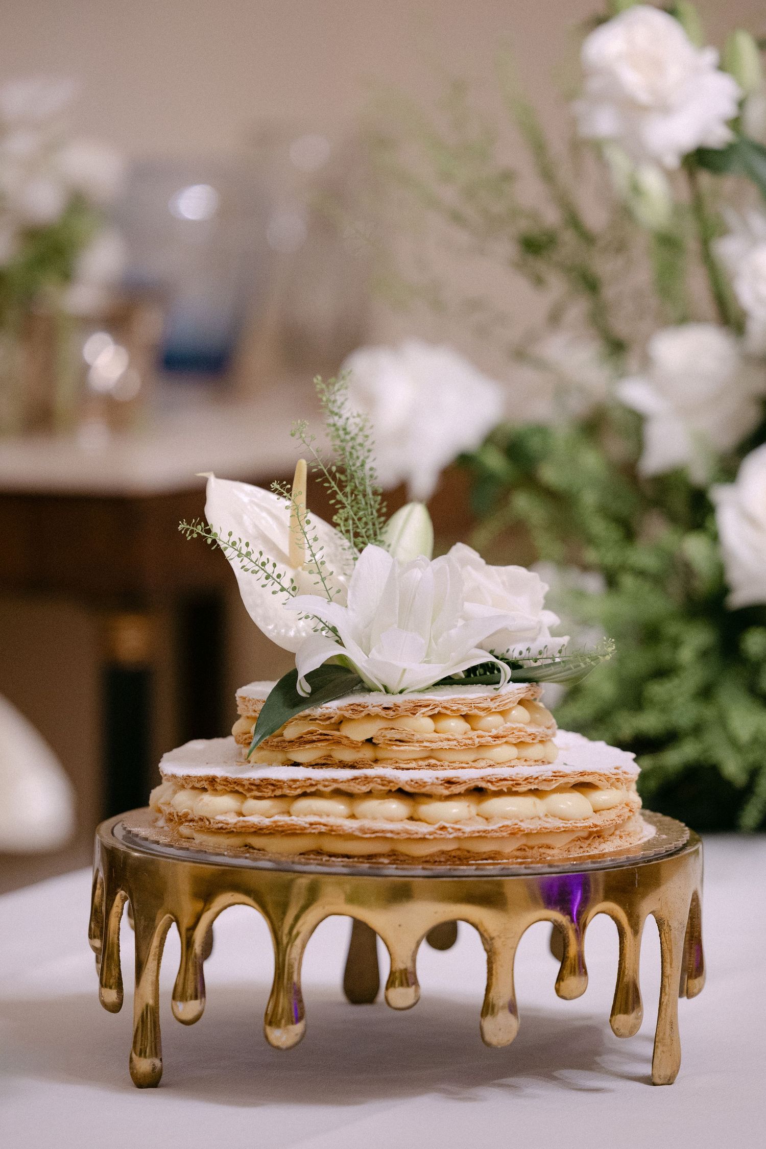 Rustic naked wedding cake topped with white lilies displayed on gold drip cake stand.