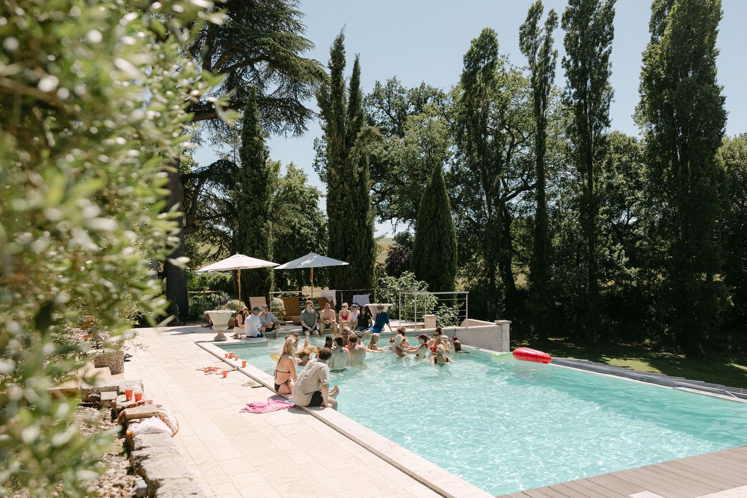 Guests enjoy a sunny outdoor pool party surrounded by tall cypress trees and lush gardens at a elegant venue.