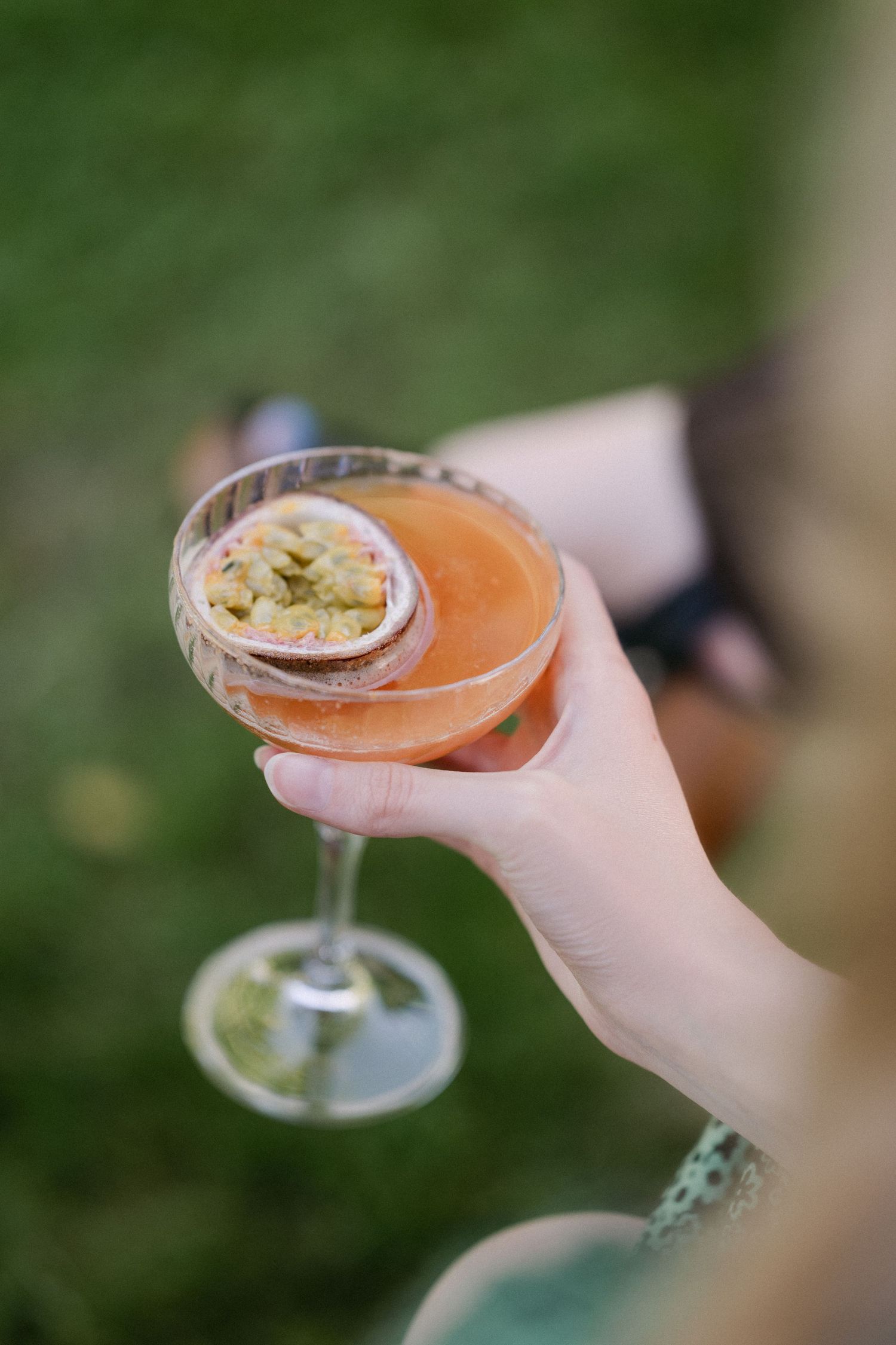 Hand holding elegant coupe glass with pink passion fruit cocktail at outdoor wedding reception.