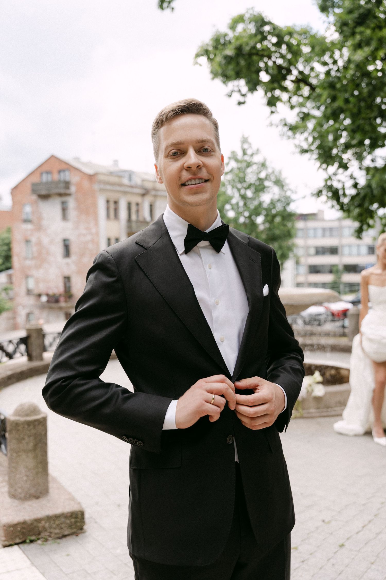 Smiling groom in a black tuxedo with bow tie buttoning his jacket outdoors in an urban setting.