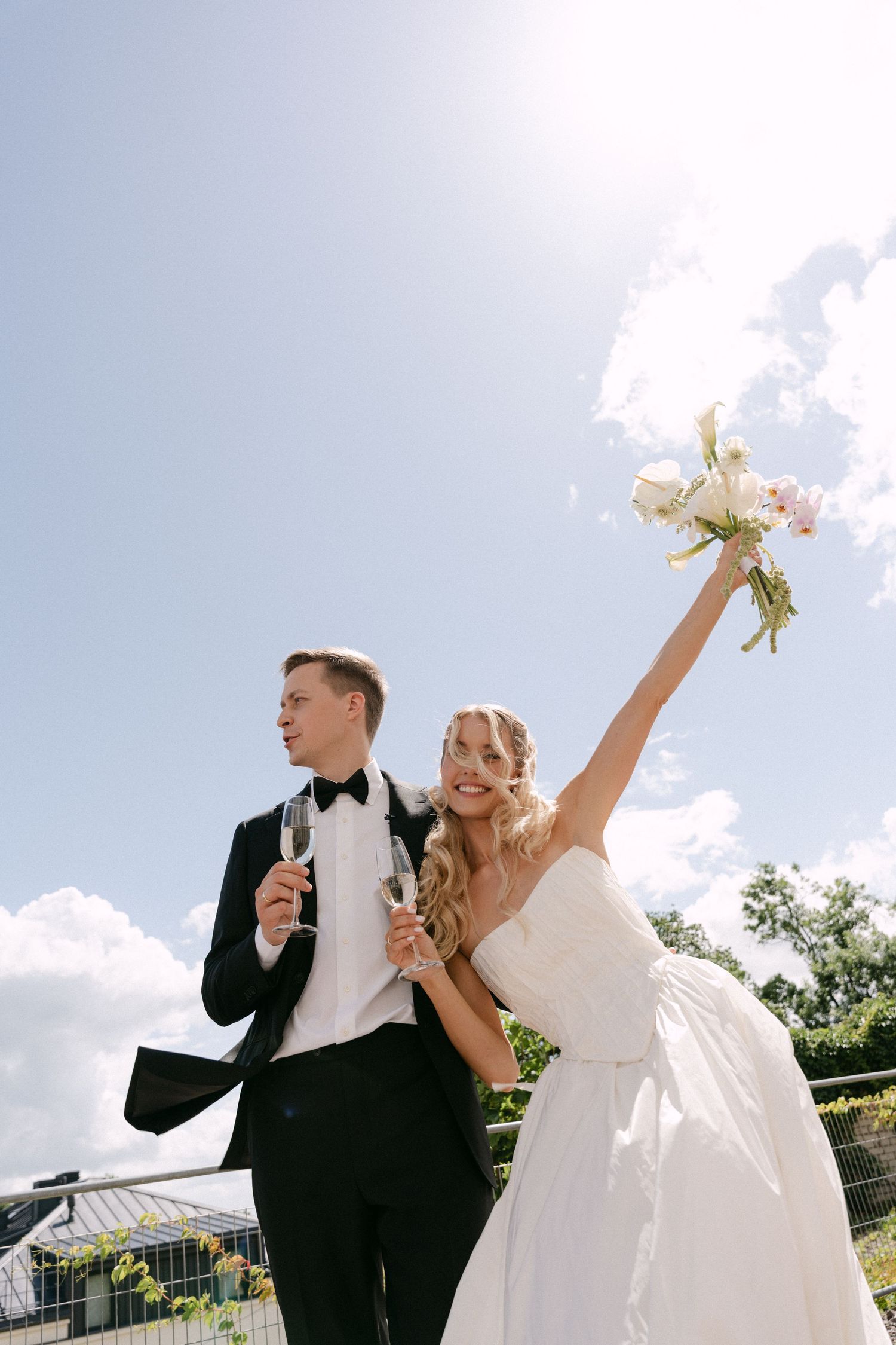 Joyful bride raises bouquet toward blue sky while groom holds champagne glass outdoors.