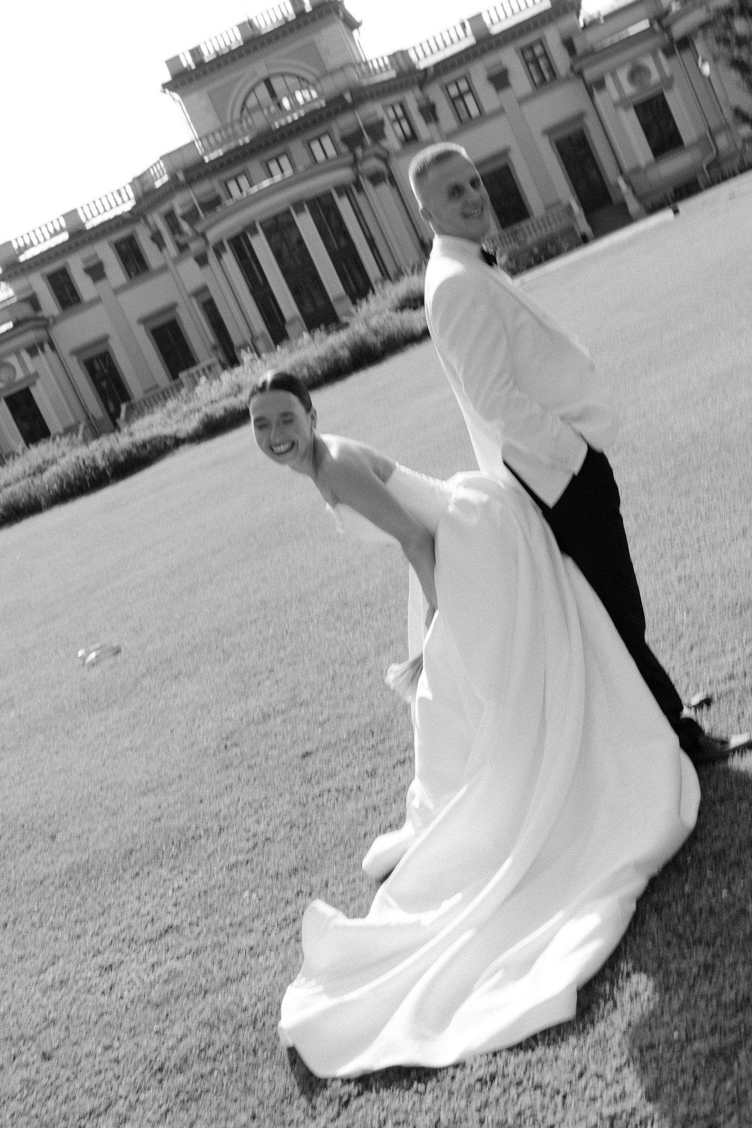 Black and white photo of joyful bride and groom running on lawn outside grand estate.