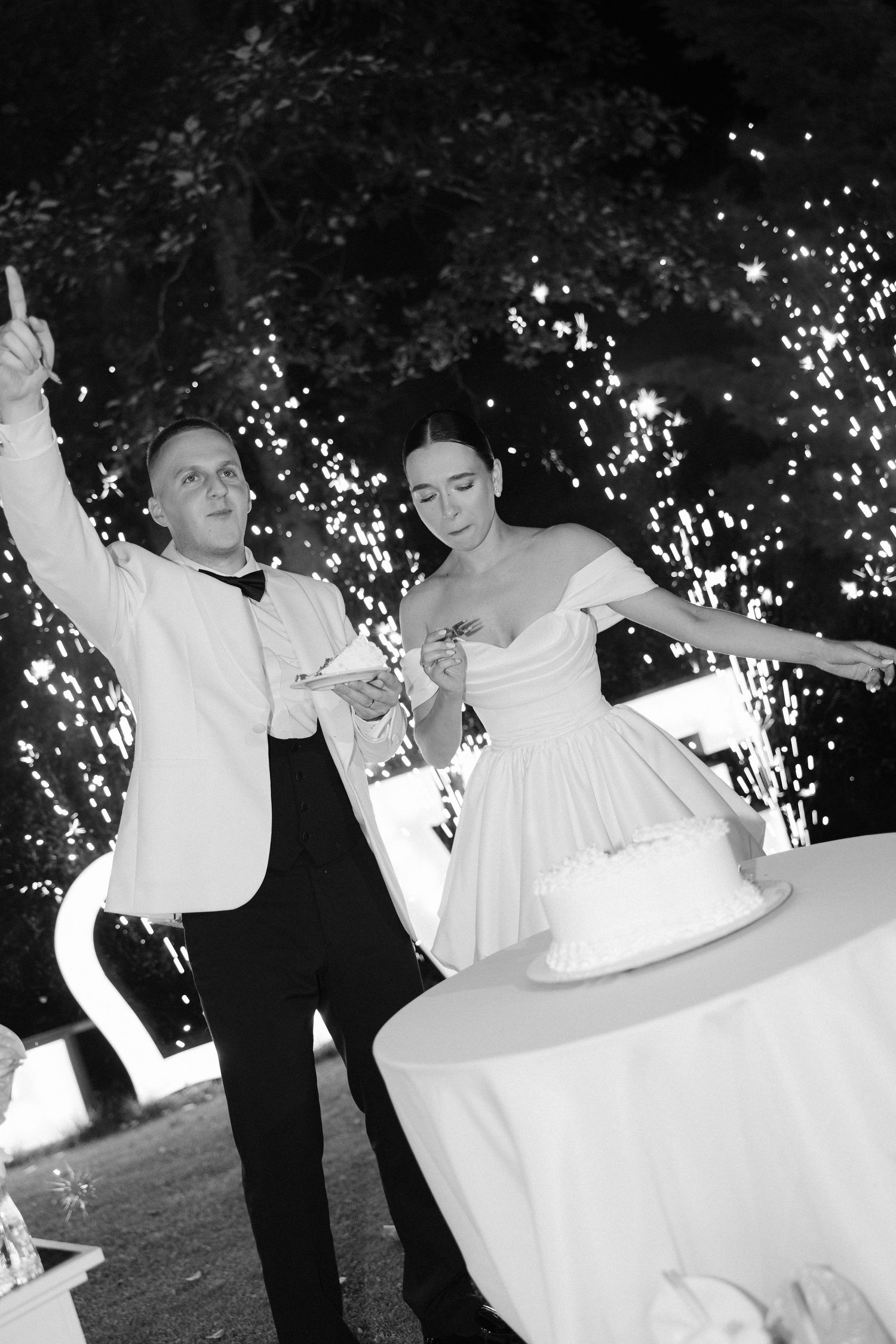 Black and white photo of joyful bride and groom cutting wedding cake amid sparkling fireworks at night reception.