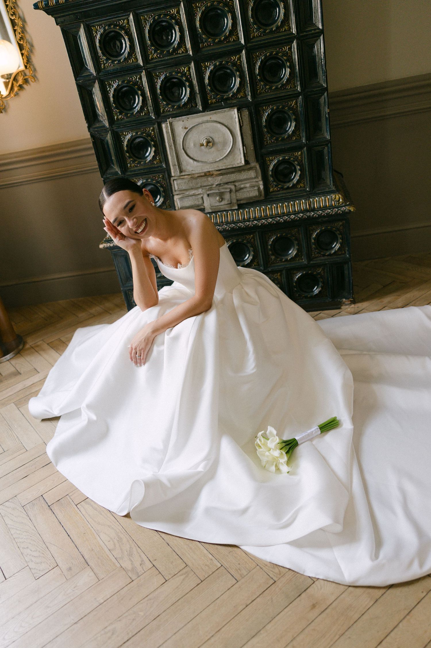 Smiling bride in white ball gown sits on herringbone floor beside antique dark tiled fireplace with calla lily bouquet.