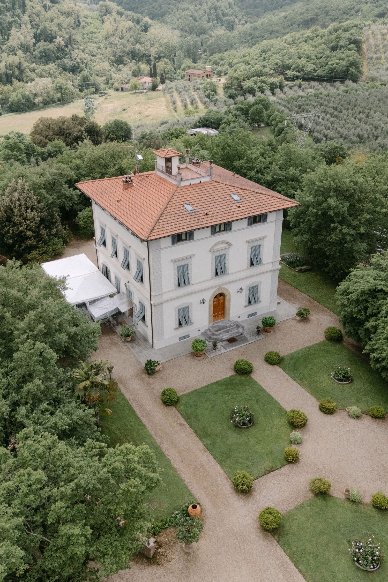 Aerial view of a grand Italian villa with terracotta roof, formal gardens, and lush Tuscan hillside surroundings.