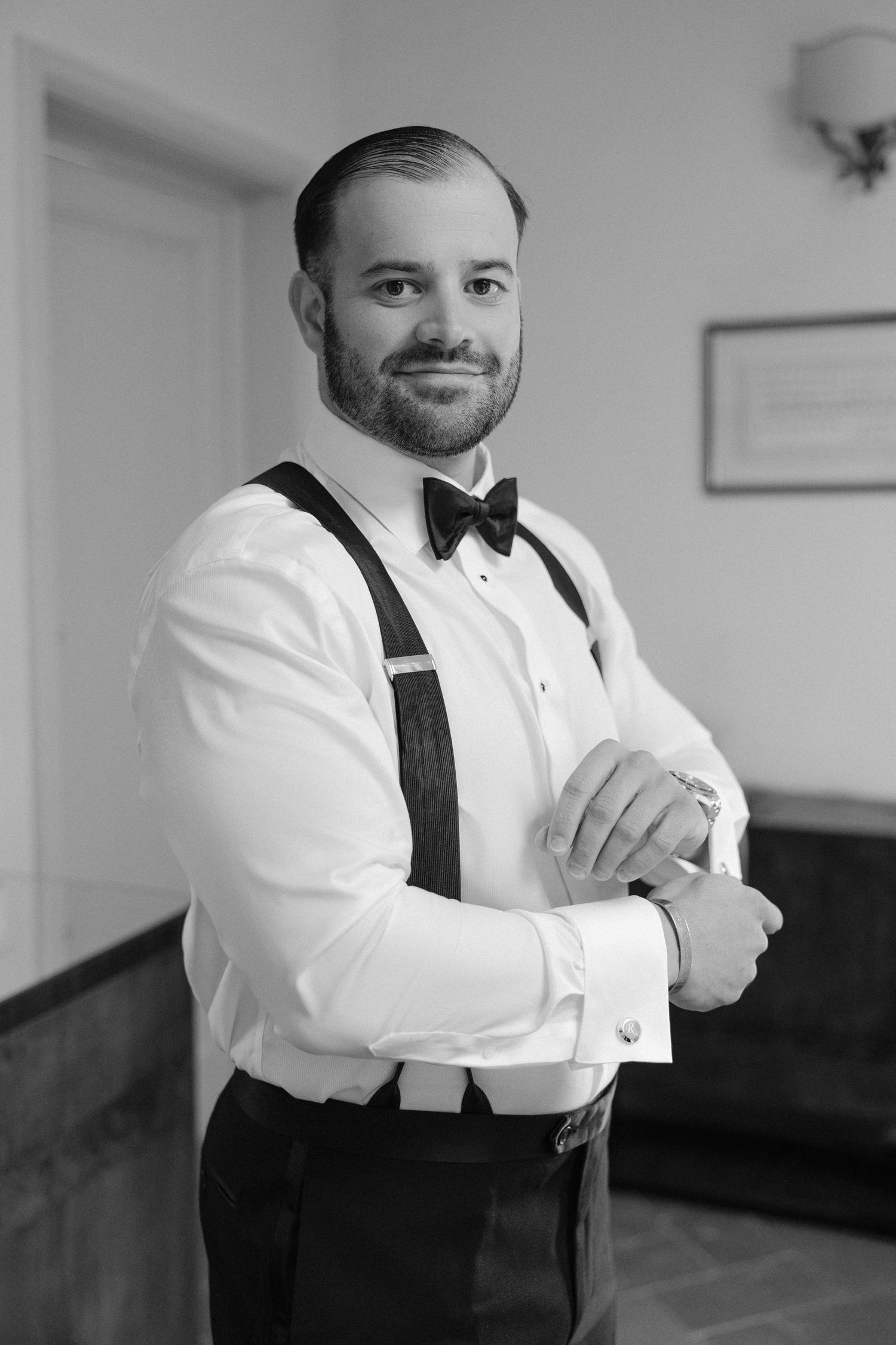Dapper groom in white shirt, suspenders, and bow tie adjusts cufflinks in black and white portrait.