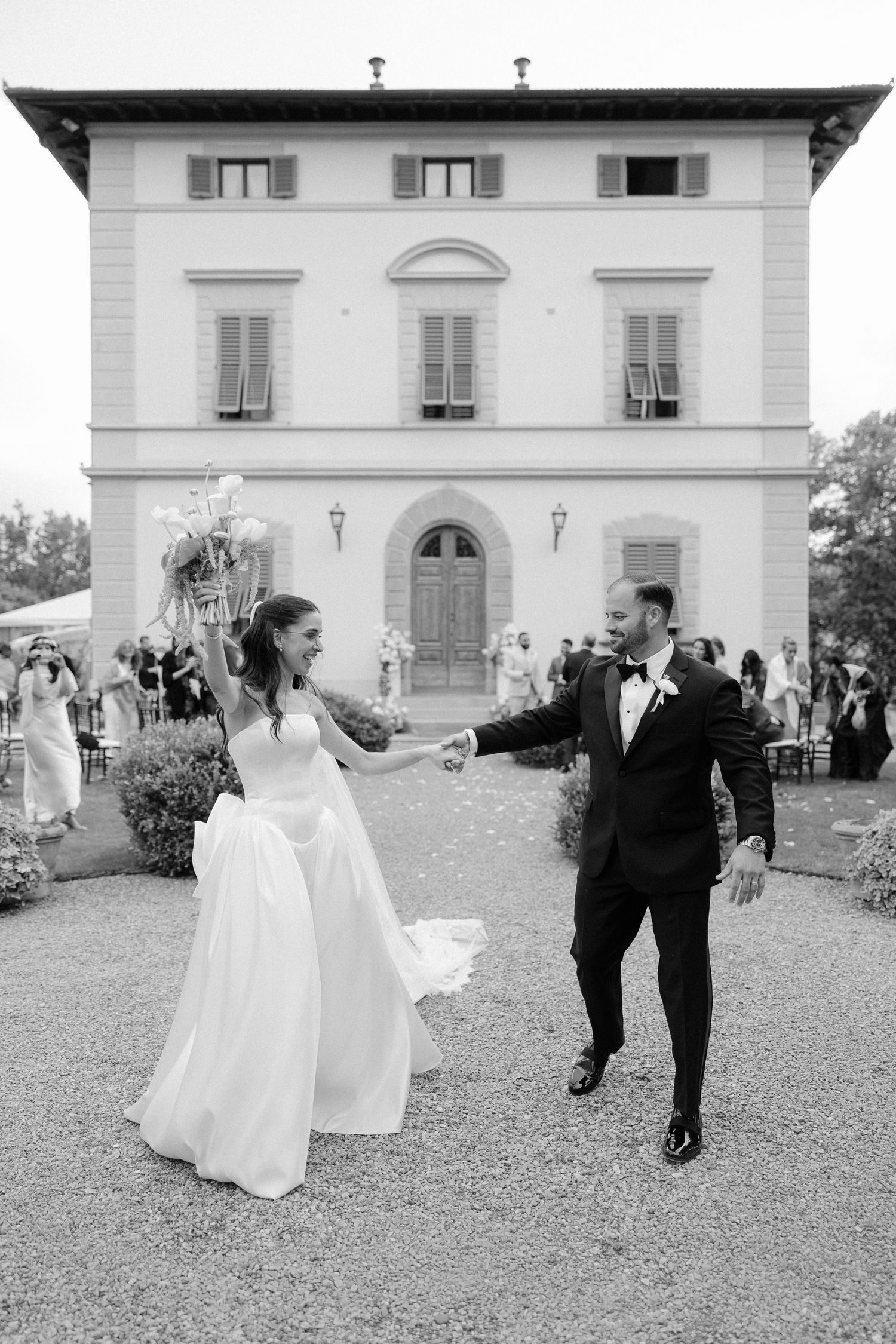 Bride and groom celebrate joyfully outside an elegant Italian villa in black and white photo.