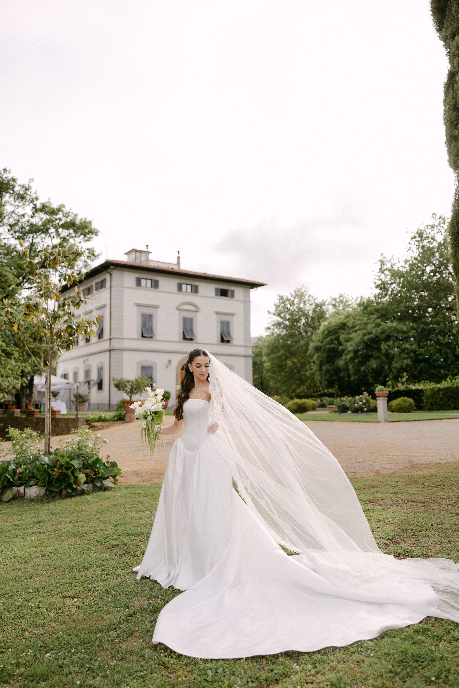 Bride in a flowing white gown with cathedral veil billowing in the breeze at a Tuscan villa.