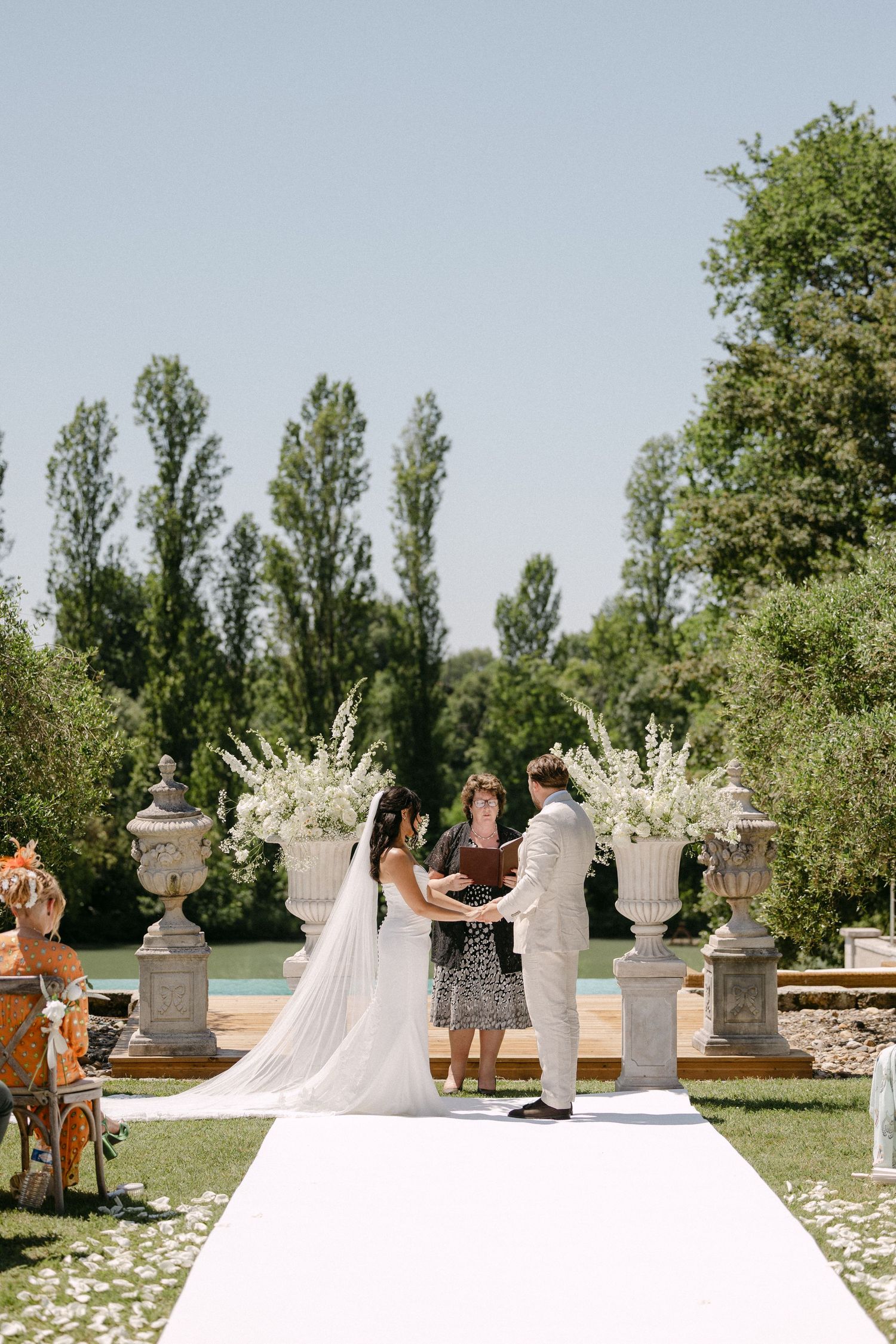 Bride and groom exchanging vows at outdoor altar with white floral arrangements and stone urns.