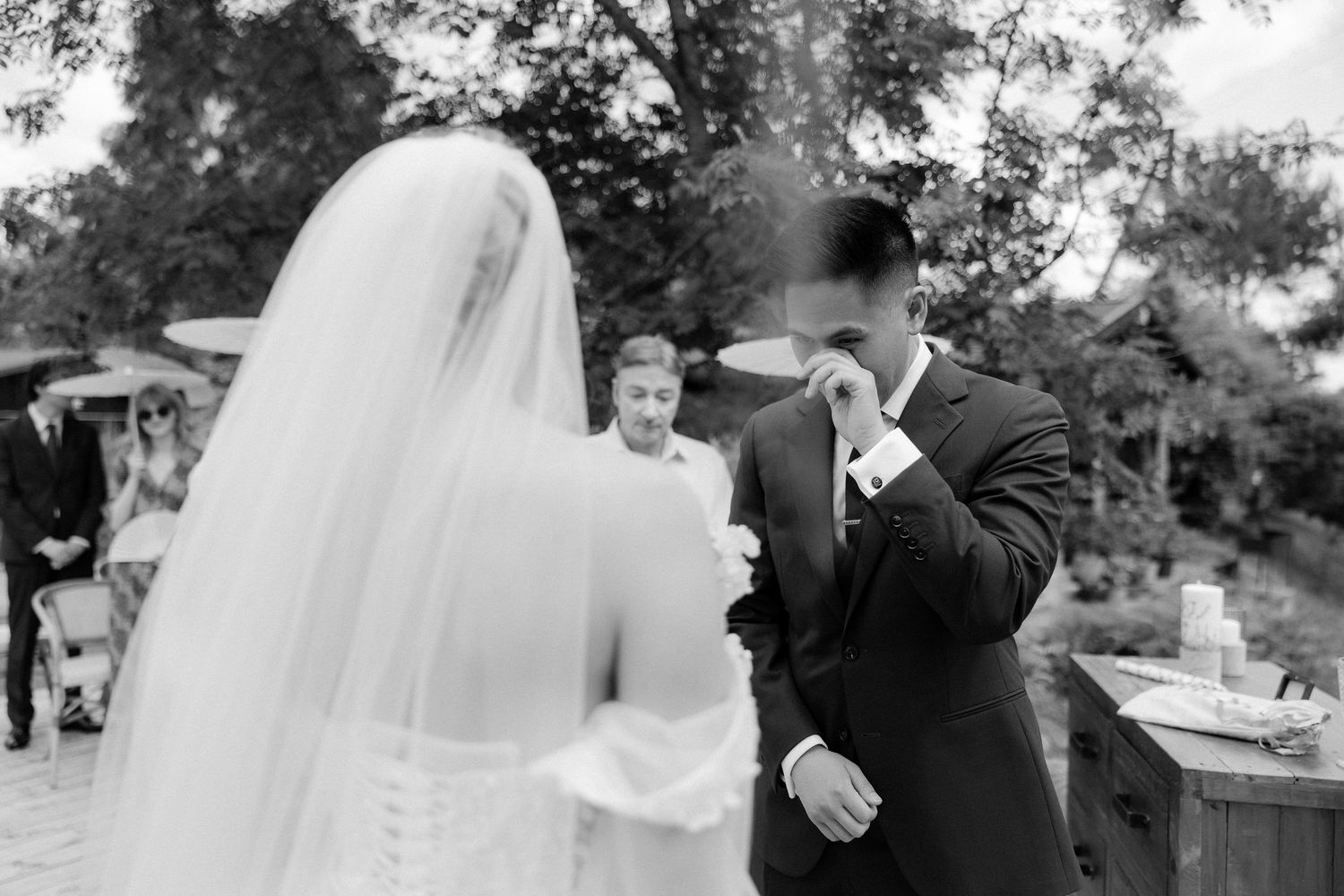 Emotional groom wiping tears as bride approaches in veil during outdoor wedding ceremony in black and white.