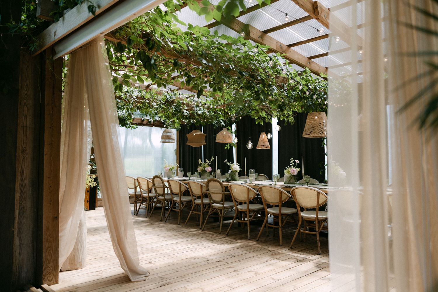 Elegant outdoor dining setup with long tables, rattan chairs, hanging lights, and lush greenery under a pergola.