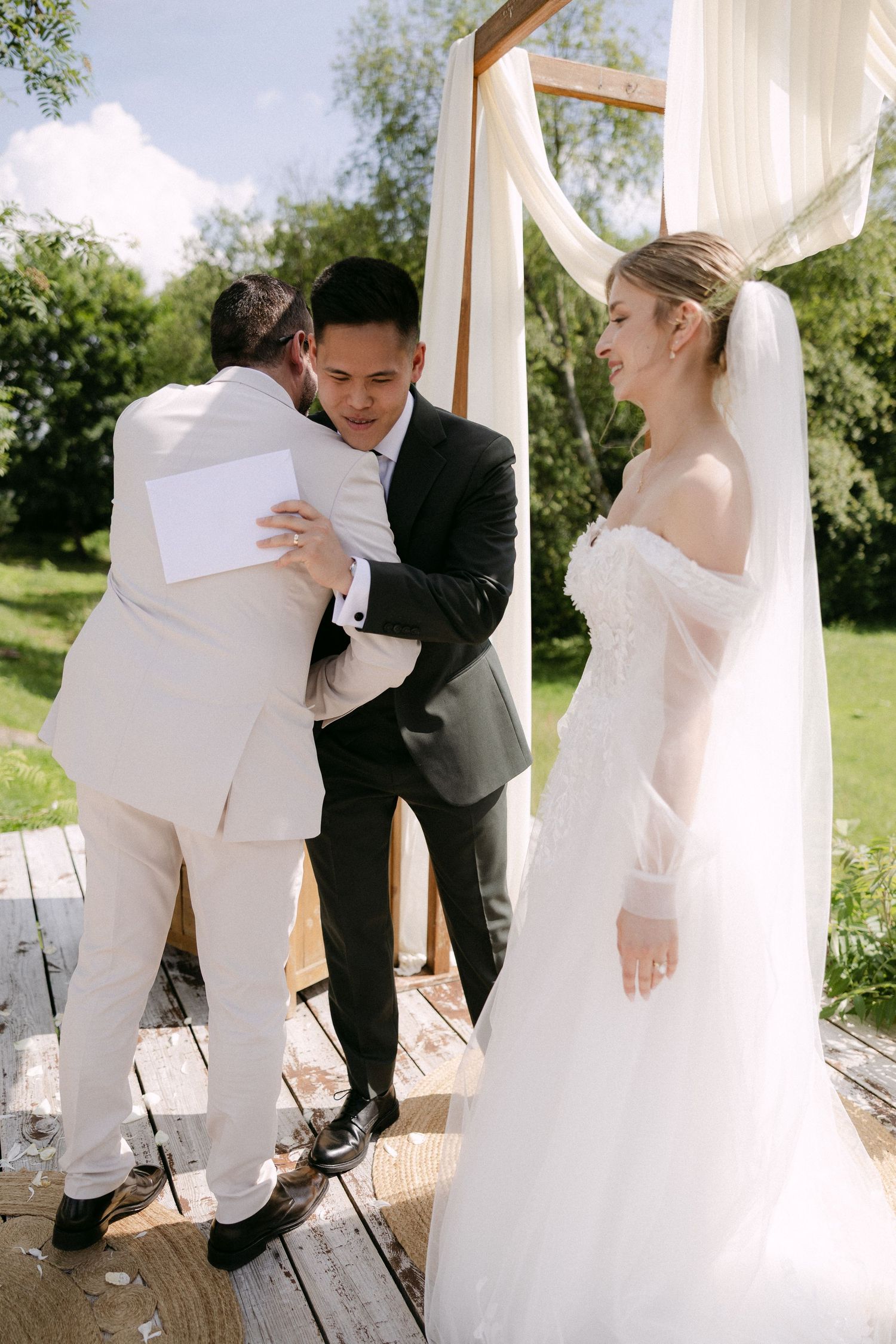 Groom hugging officiant while bride smiles during outdoor wedding ceremony.