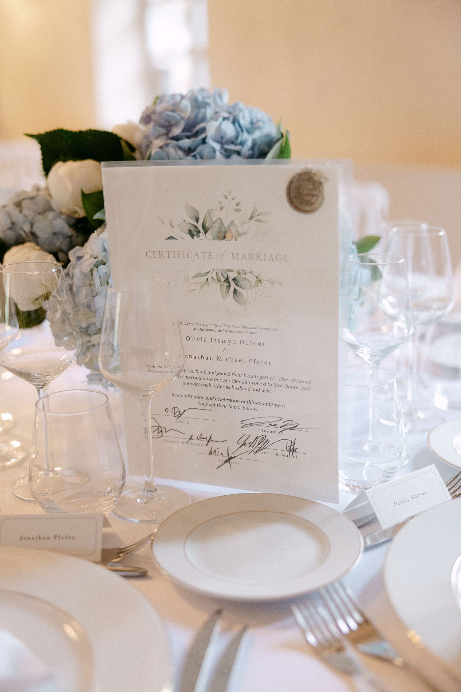 Elegant wedding table setting with certificate of marriage, blue hydrangeas, white plates, and crystal glassware.