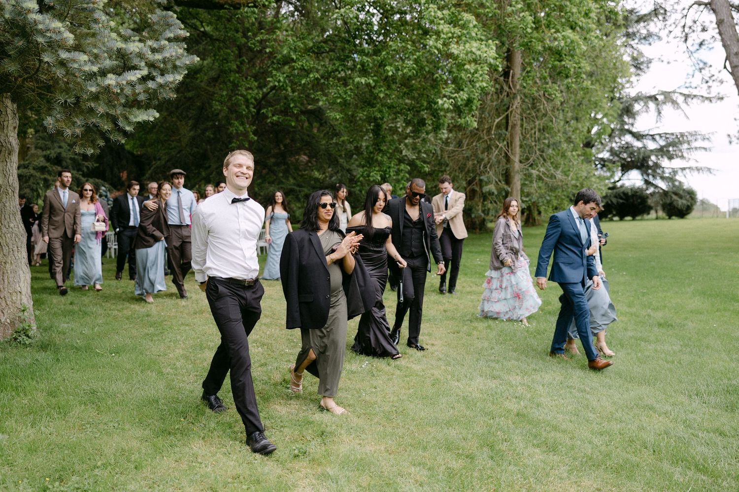 Wedding guests in formal attire walking across a green lawn surrounded by tall trees at an outdoor reception.