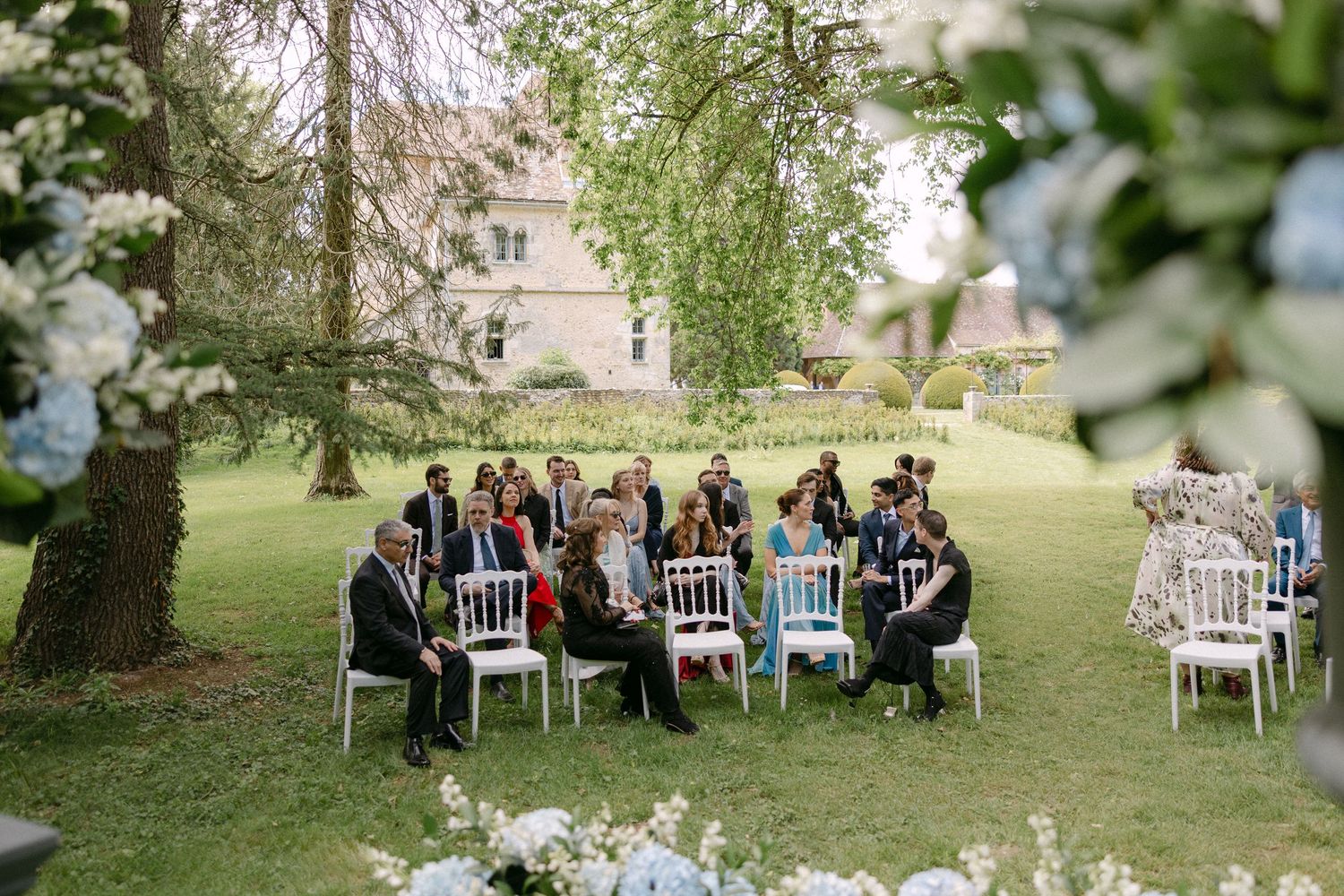 Guests seated on white chairs for an outdoor wedding ceremony on a lush green lawn surrounded by blooming trees.