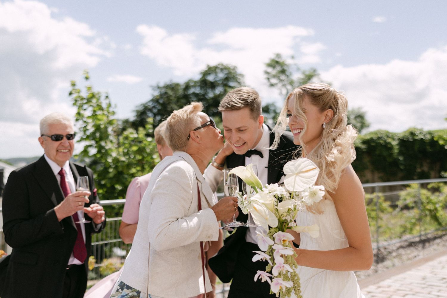 Joyful bride and groom receiving kiss from elderly guest outdoors at sunny wedding celebration.