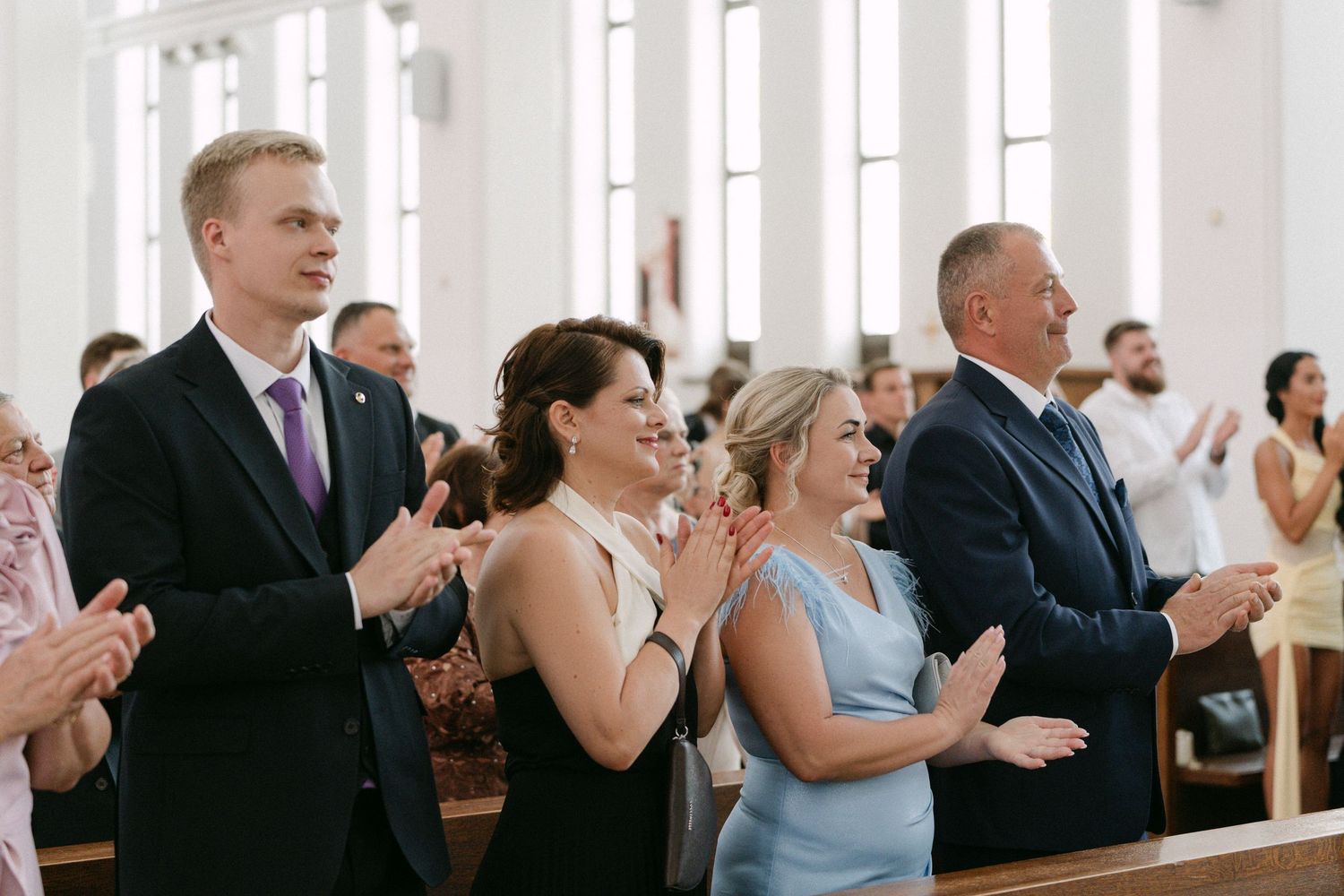 Wedding guests clapping and smiling in a white church during a ceremony.