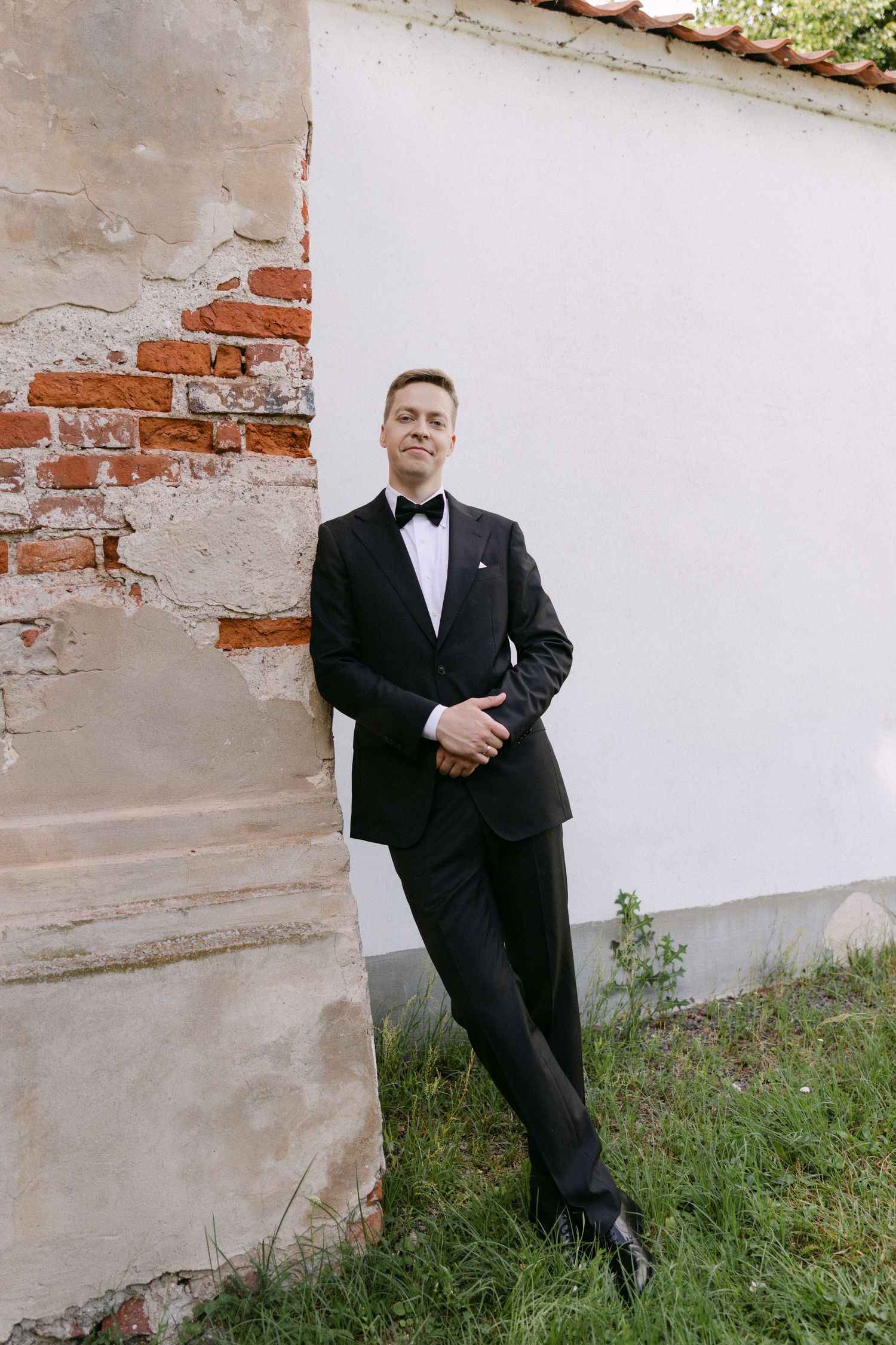 Dapper groom in a black tuxedo leans against a rustic brick and white wall outdoors.
