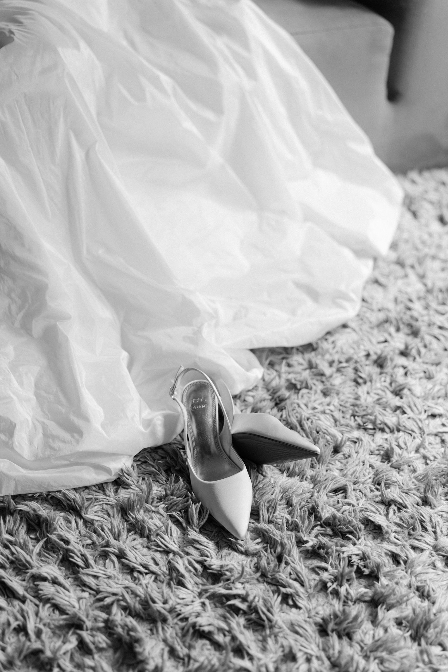 Black and white photo of elegant bridal heels on carpet beside flowing white wedding dress.