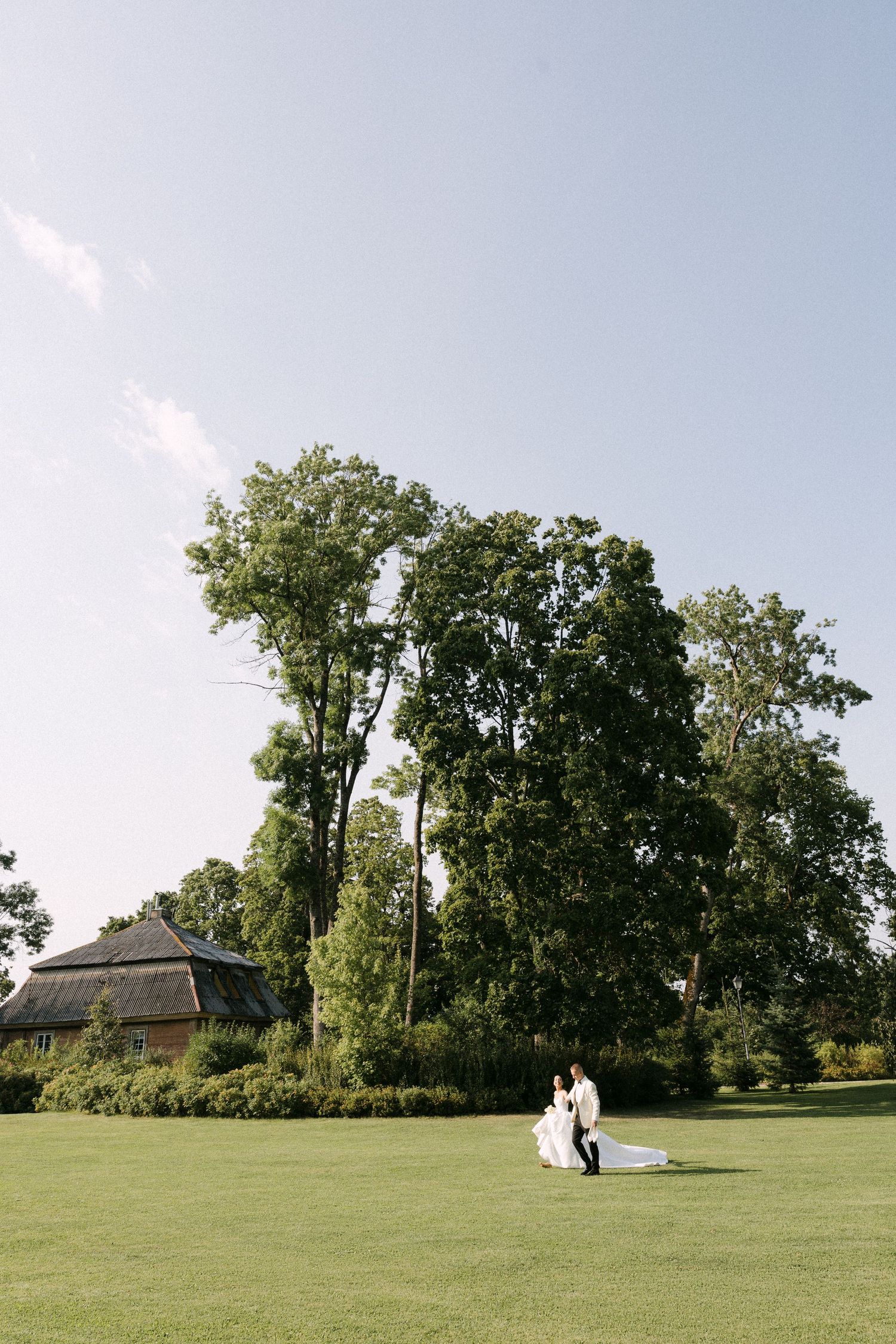 Bride and groom embrace on lush green lawn surrounded by tall trees and rustic barn in summer sunlight.