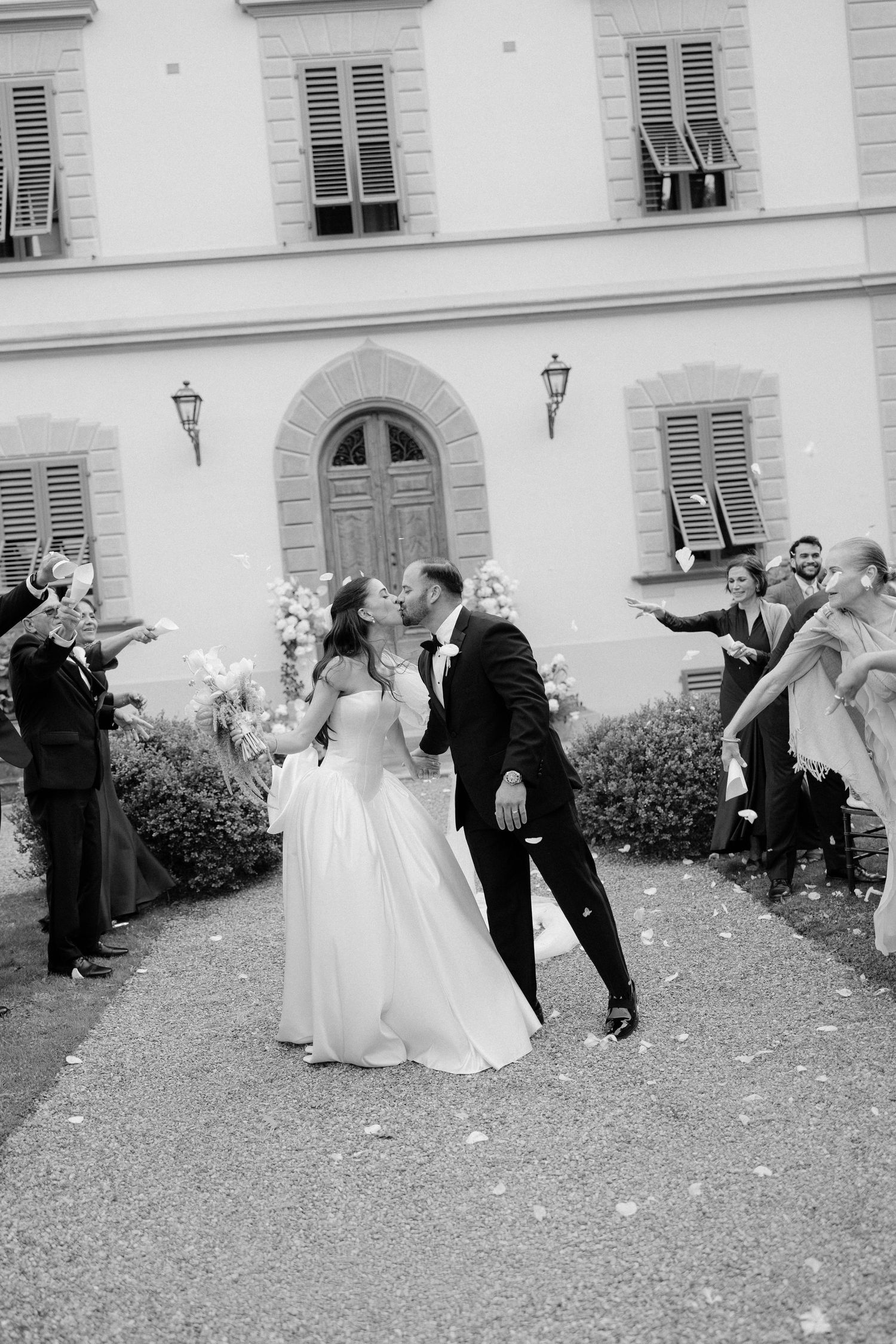 Black and white photo of bride and groom kissing during confetti exit at elegant Italian villa wedding.