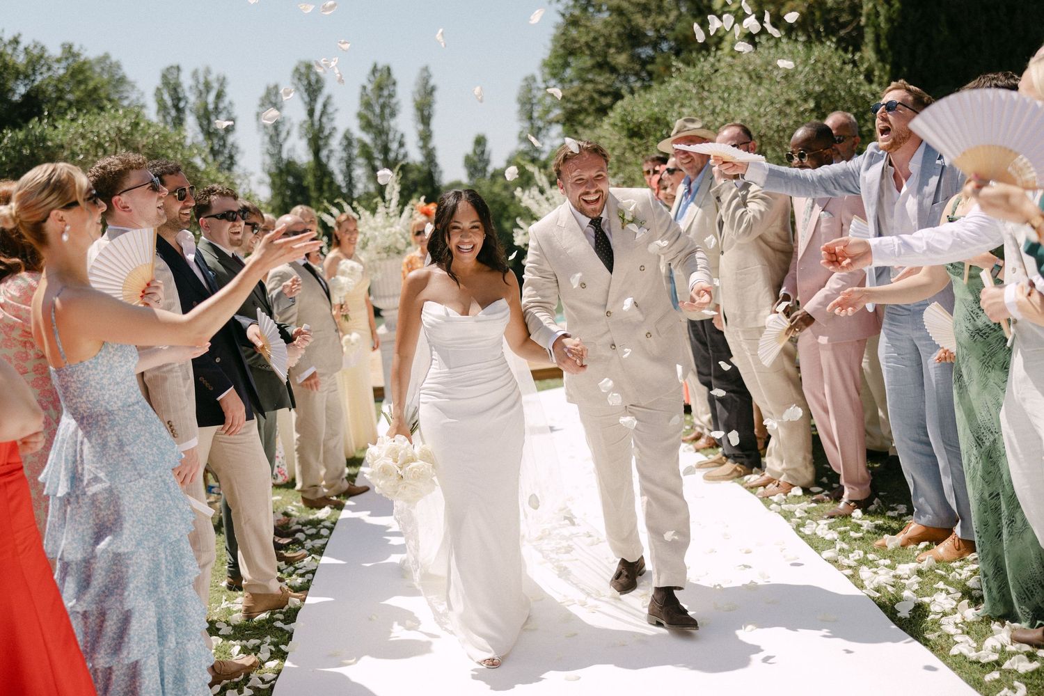 Bride and groom walk down aisle as guests toss confetti at outdoor wedding ceremony.